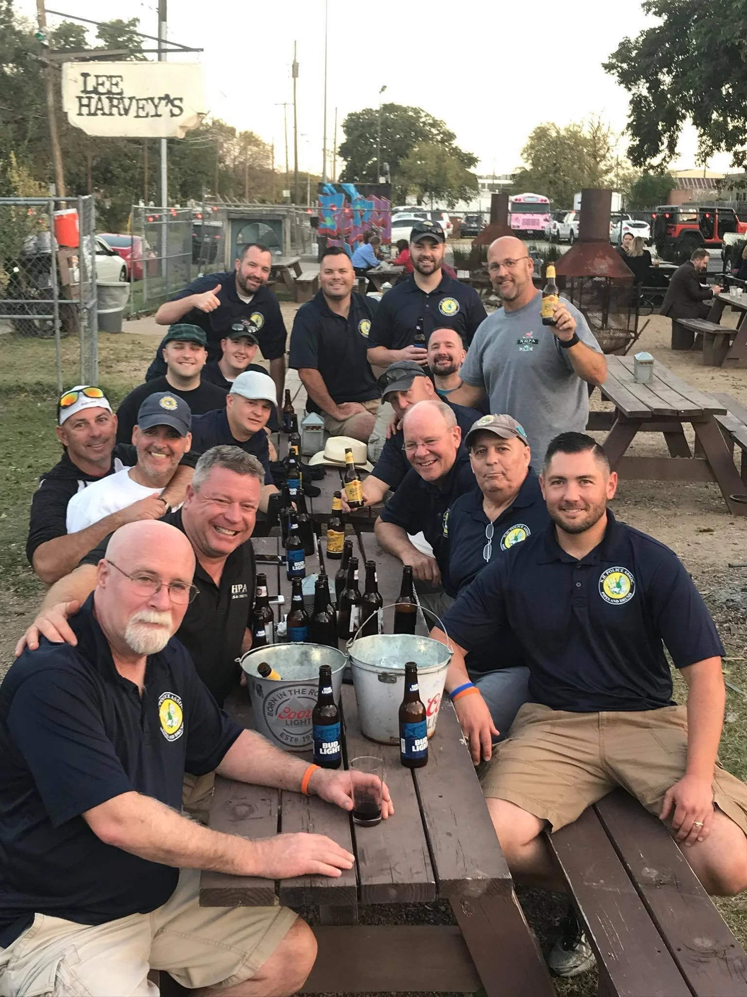 Group of men gathered at a picnic table outdoors, holding drinks and smiling, with a sign reading 'Lee Harvey's' in the background.