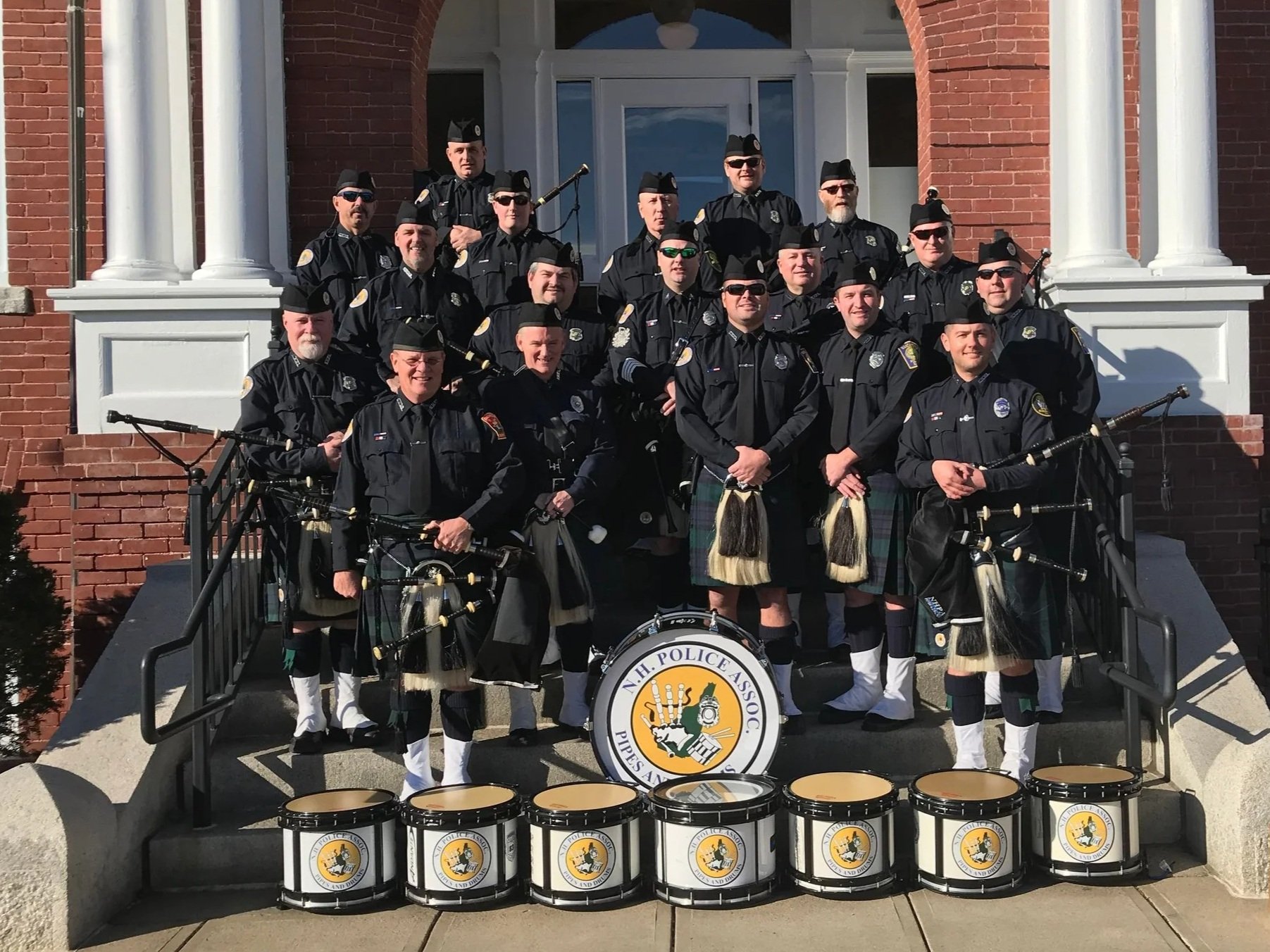 Group of police officers in uniform, some playing bagpipes, standing on the steps of a brick building with white columns. There are drums with a police association logo in front of them.