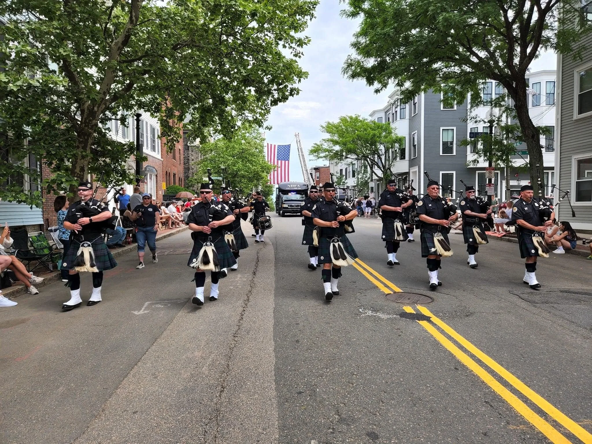 A parade with a marching band of people dressed in traditional Scottish kilts, playing bagpipes and drums, walking down a street lined with spectators and trees. An American flag and a fire truck are visible in the background.