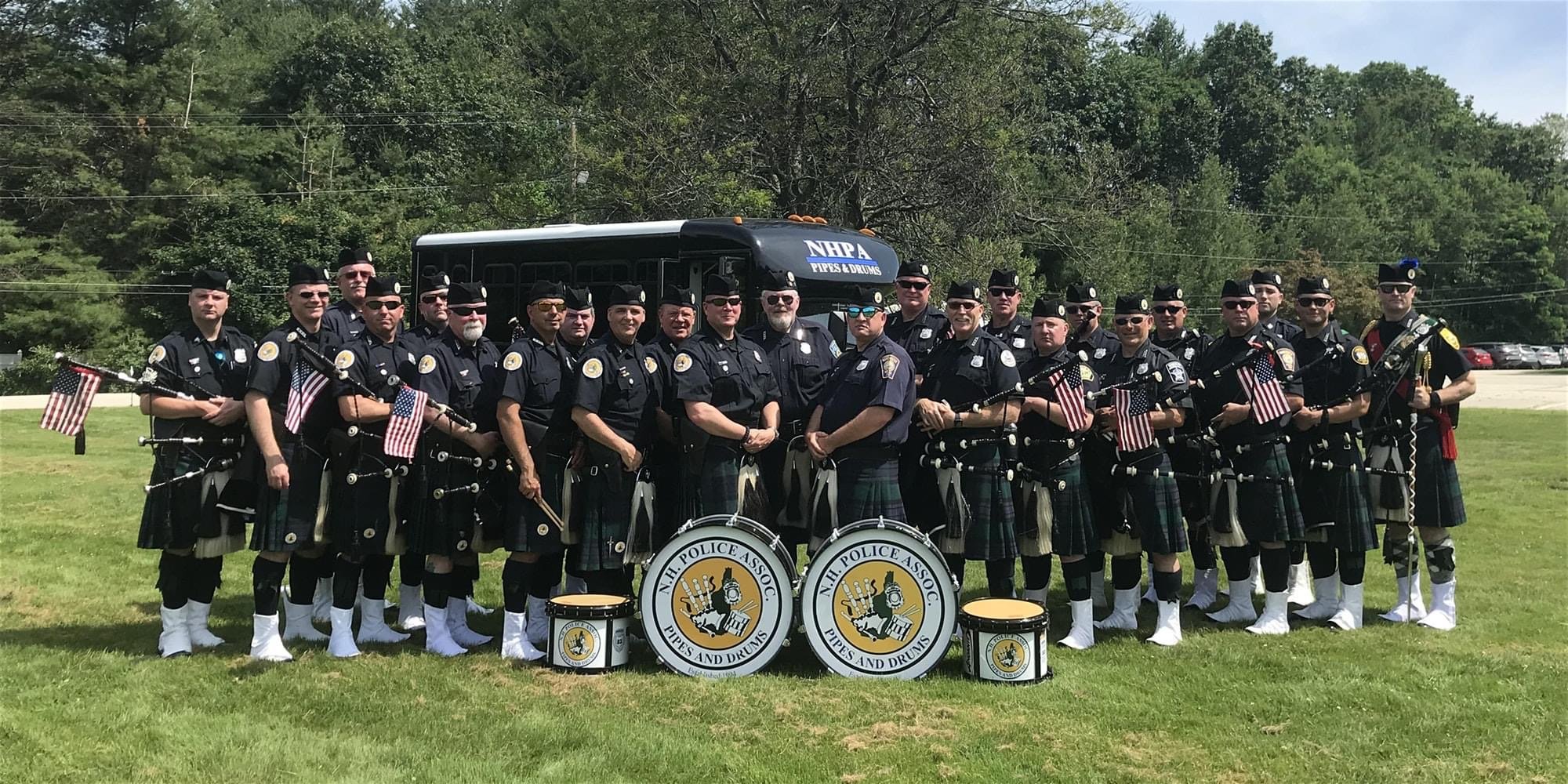 A group of police officers dressed in uniforms and kilts standing outdoors on a grassy field. They are holding bagpipes and American flags. There are drums with the sign "N.H. Police Assoc. Pipes and Drums" in front of them, and a bus with a sign "NH