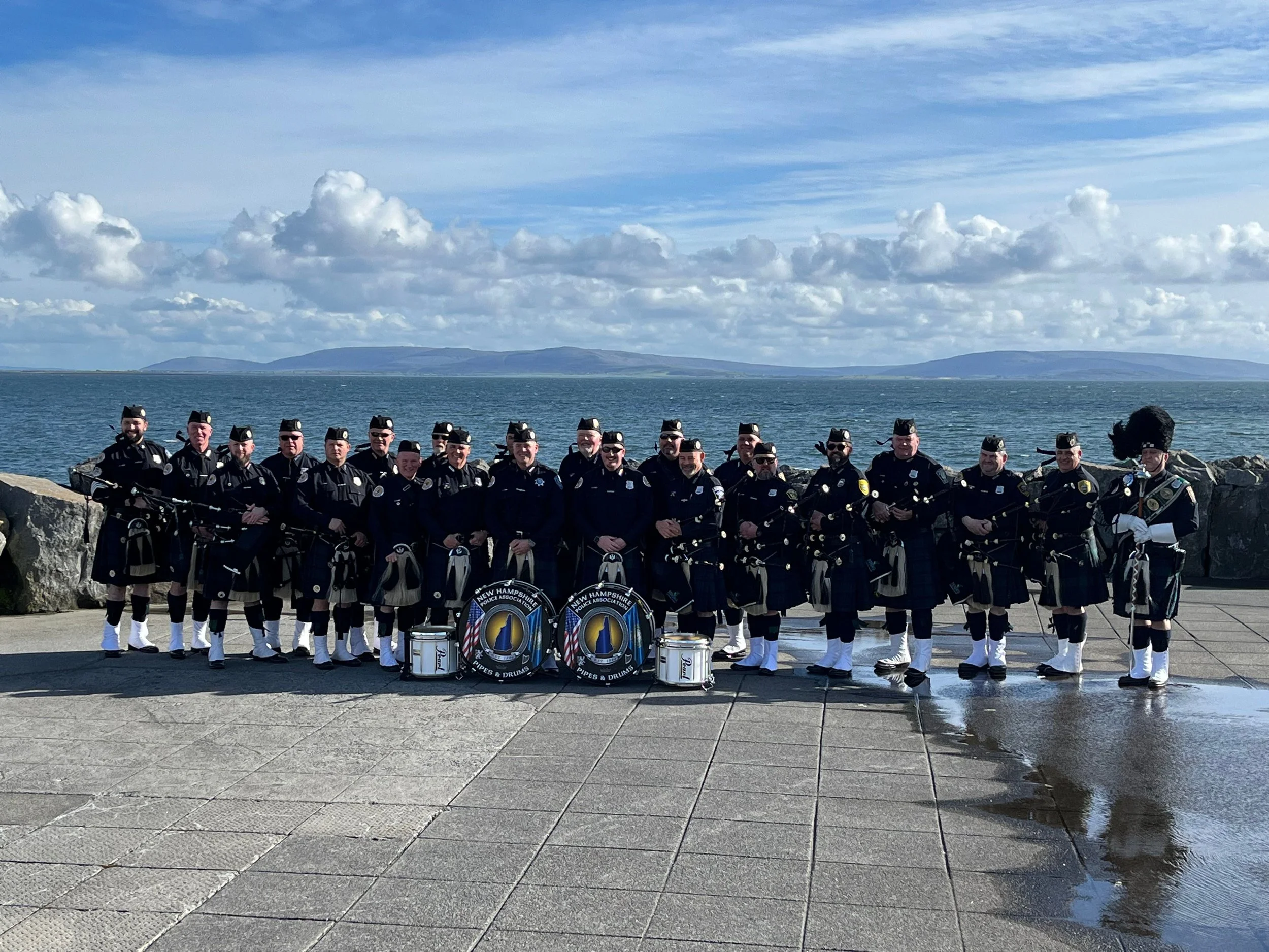 A group of musicians dressed in traditional Scottish uniforms, including kilts, standing by the water with musical instruments, in front of a scenic ocean view with distant hills and a partly cloudy sky.