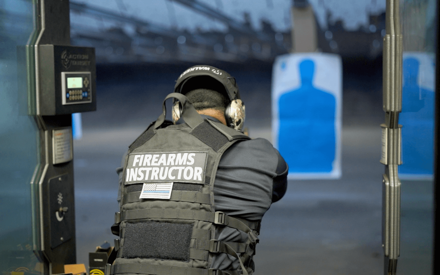 A firearm instructor wearing protective gear and a vest labeled 'FIREARMS INSTRUCTOR' aims at a shooting target at an indoor shooting range.