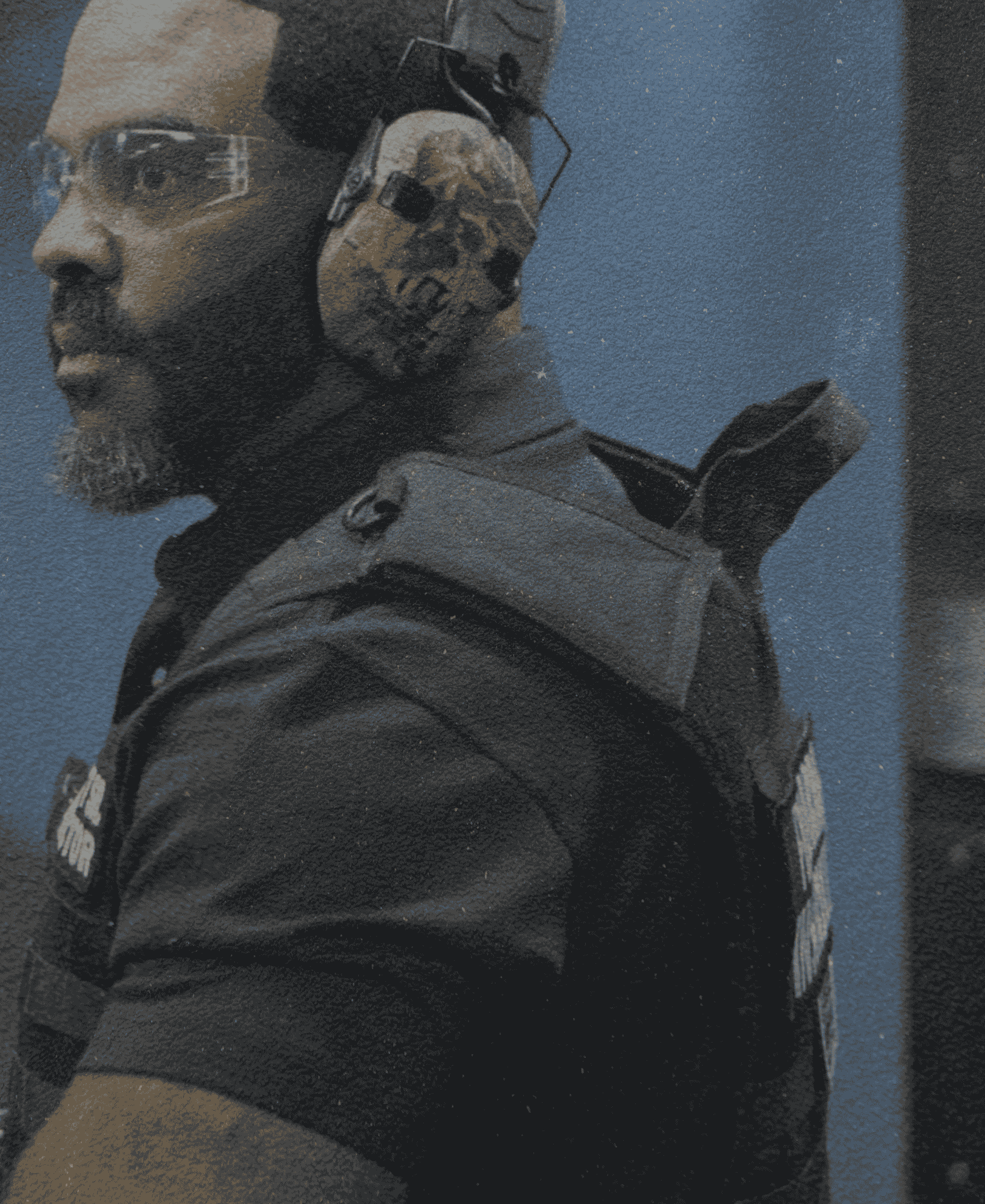 Close-up of a person in tactical gear wearing a headset with a skull emblem on the side, against a blue wall.