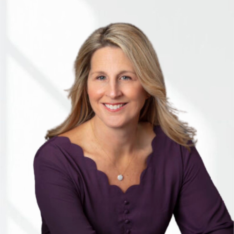 A woman with shoulder-length blonde hair, smiling, wearing a purple top with scalloped edges, and a small necklace, against a plain, light background.