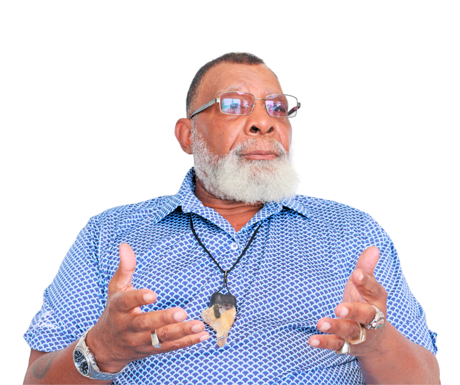An older man with a white beard wearing sunglasses, a blue patterned shirt, and a necklace with a large stone pendant. He is gesturing with his hands.