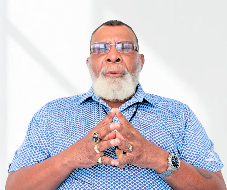A man with a gray beard and glasses wearing a blue patterned shirt, holding his hands together with fingers interlocked and looking at the camera.
