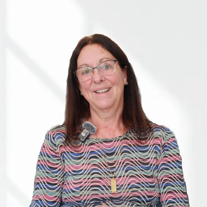 Smiling woman wearing glasses, patterned multicolored top, with microphone and gold necklace, in front of white background.