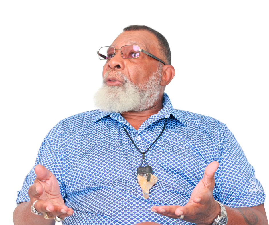 An older man with glasses, a white beard and mustache, wearing a blue patterned shirt, a necklace with a large stone pendant, and a watch, gesturing with his hands while speaking.