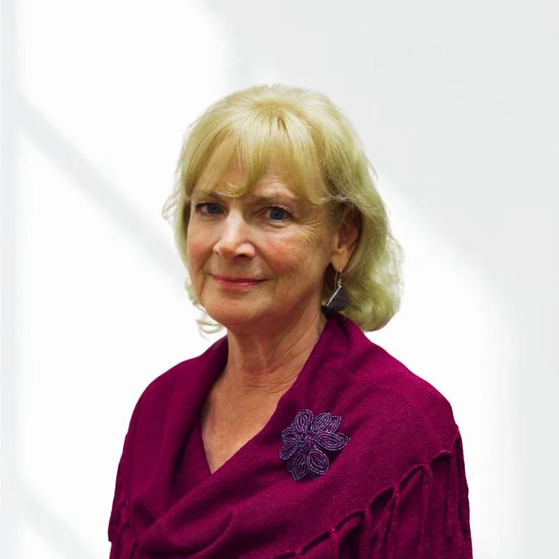 An older woman with blonde hair, wearing a purple top with a floral embellishment, standing against a white background.