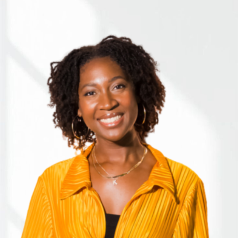 Portrait of a woman with curly hair smiling, wearing a yellow blouse with a necklace and hoop earrings, against a plain background.