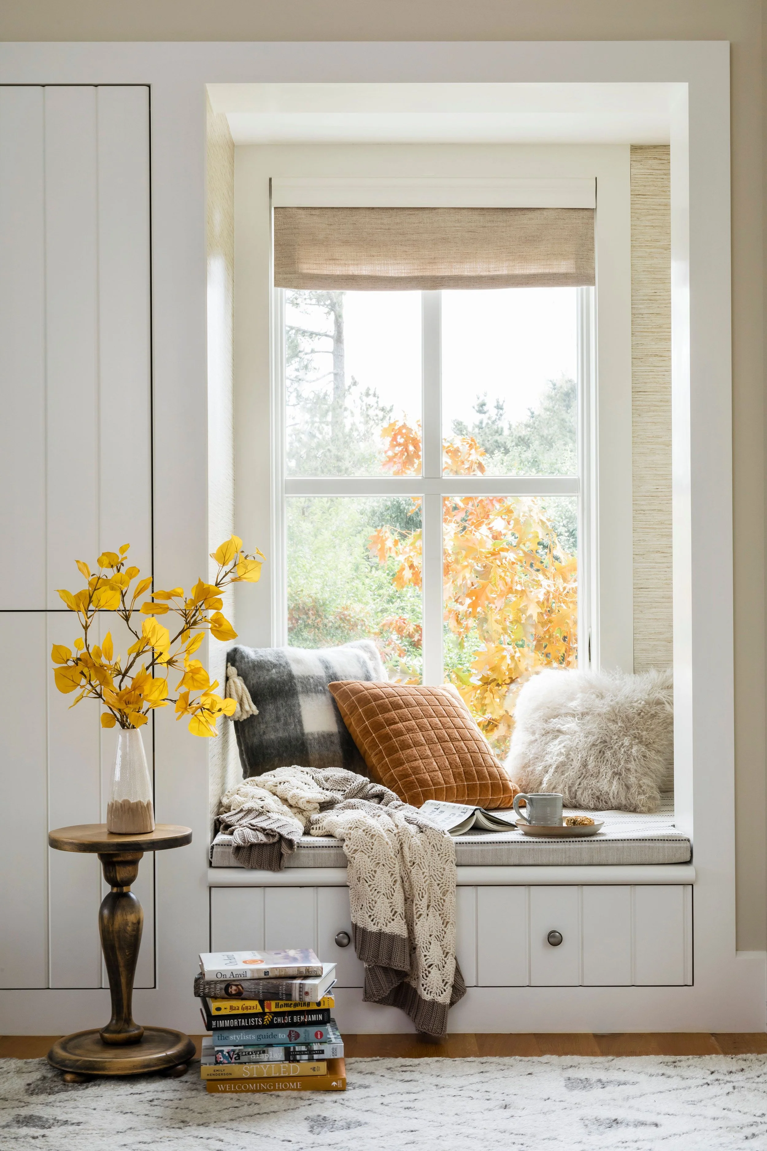 Cozy window nook with cushions, throw blankets, a cup, and an open book, decorated with a vase of yellow flowers and a stack of books in front, overlooking autumn trees outside.