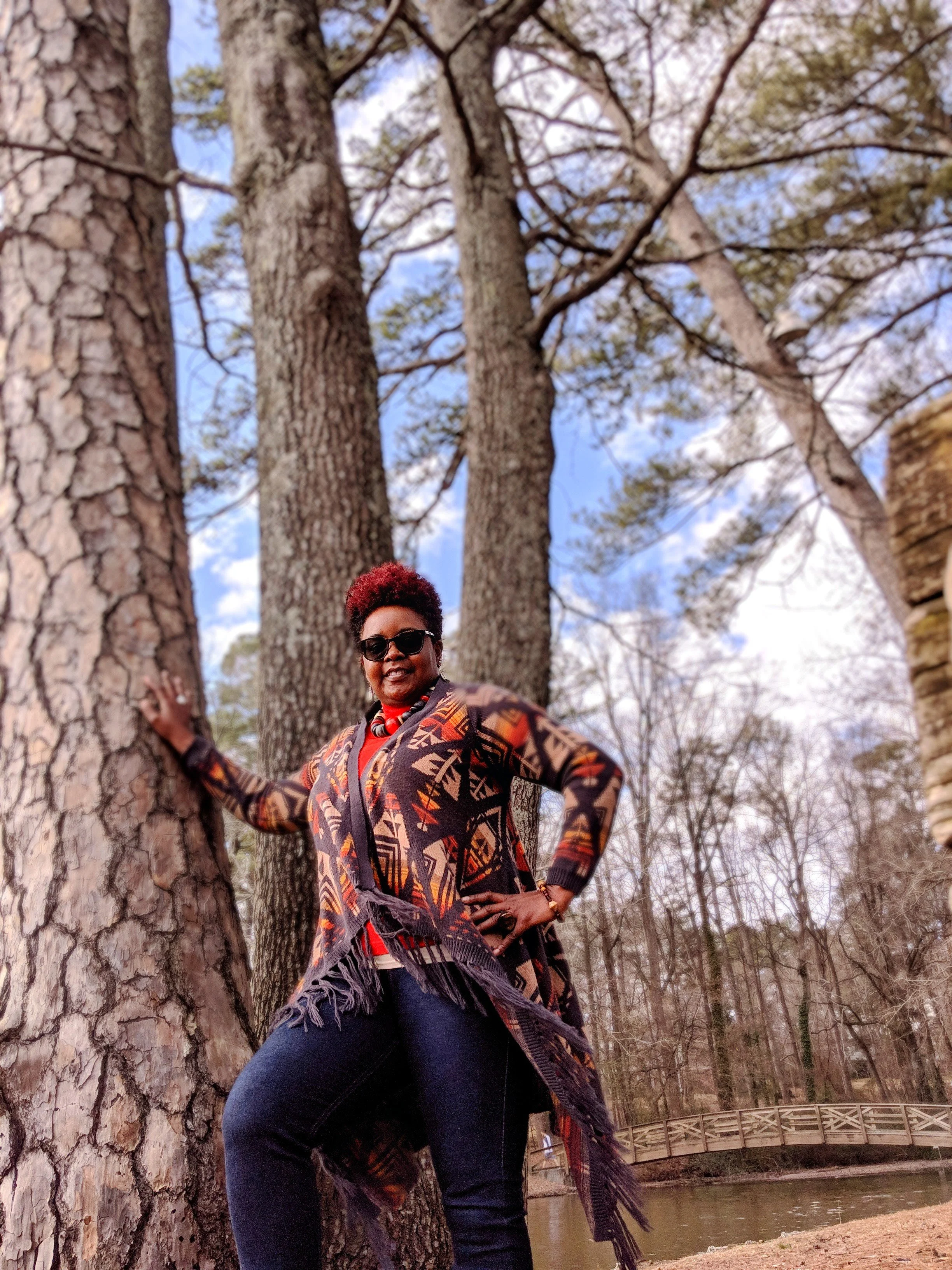 Woman standing outdoors among large trees by a park pond, wearing a colorful patterned shawl, sunglasses, and smiling.