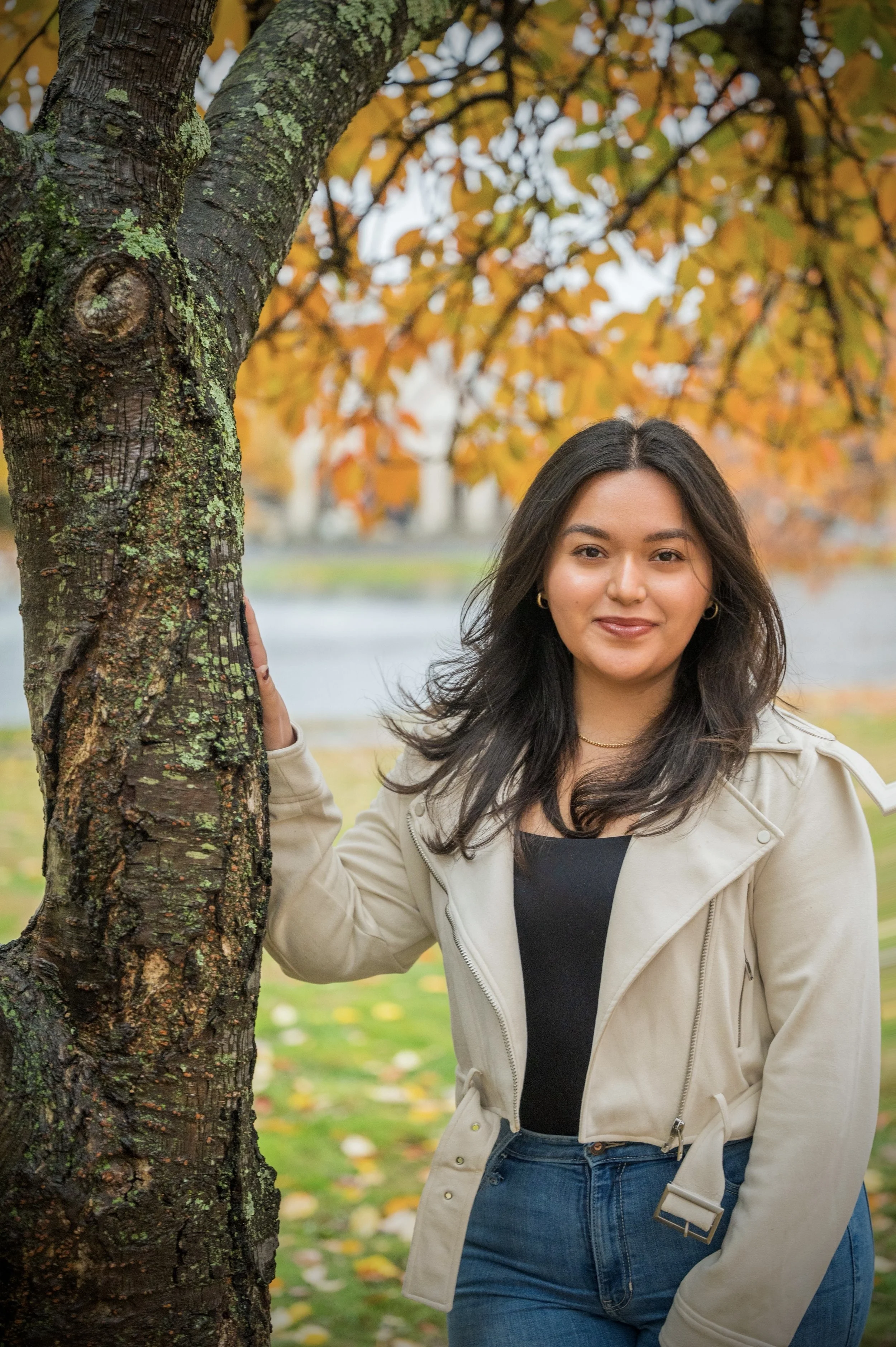 A woman with dark hair wearing a beige jacket and blue jeans, standing outdoors next to a tree with orange and green leaves in autumn.