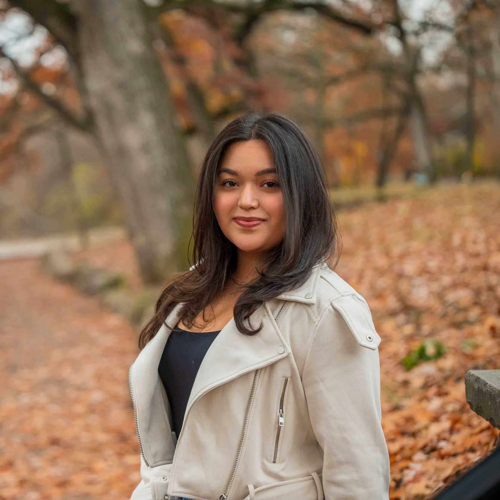 A woman with long dark hair wearing a beige jacket and black top, standing outdoors in a park with autumn leaves on the ground and trees in the background.