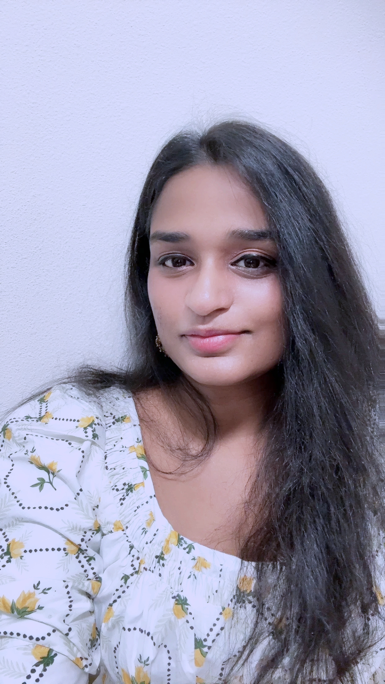 Close-up of a young woman with dark hair wearing a white top with yellow flowers, looking at the camera with a slight smile, against a plain white wall background.