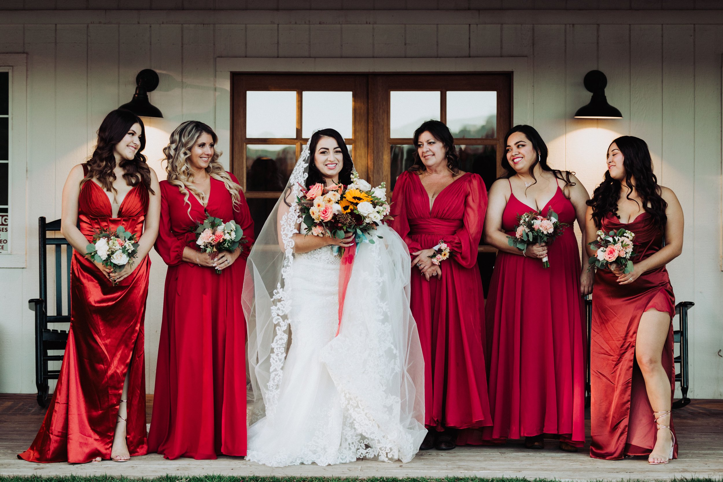 Bride and six bridesmaids dressed in red gowns, standing outside a building with wooden doors, holding bouquets of flowers.