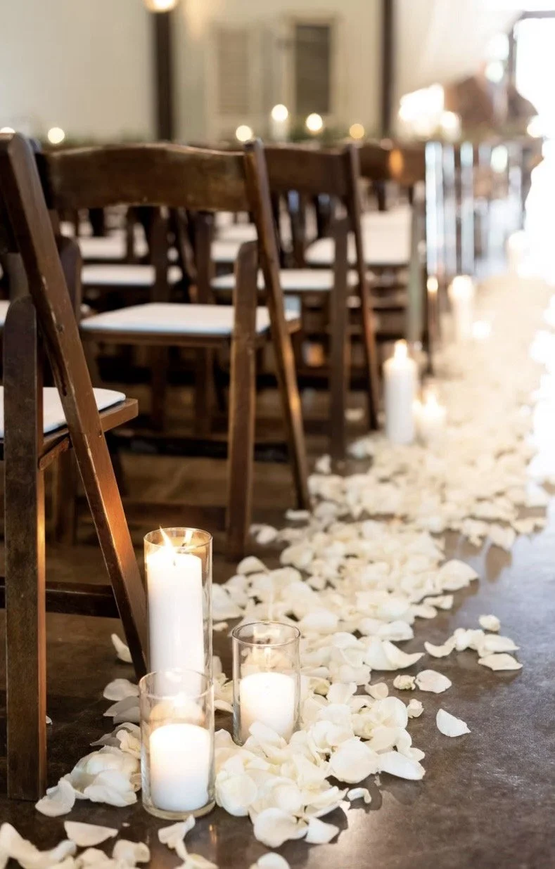 A wedding aisle decorated with white flower petals and candles in glass holders.