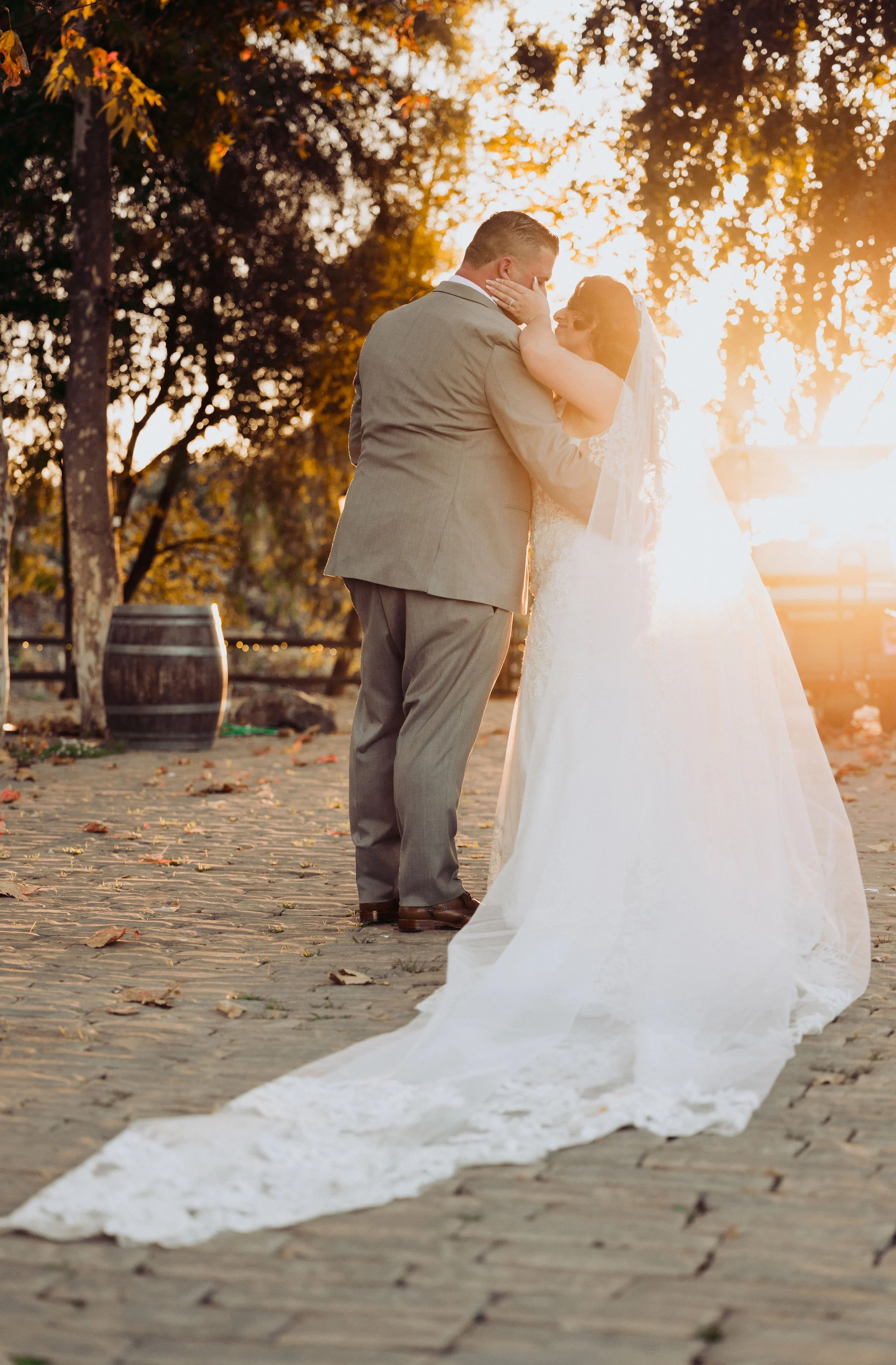 A bride and groom sharing a kiss during their wedding at sunset, outdoors with trees and fallen leaves.