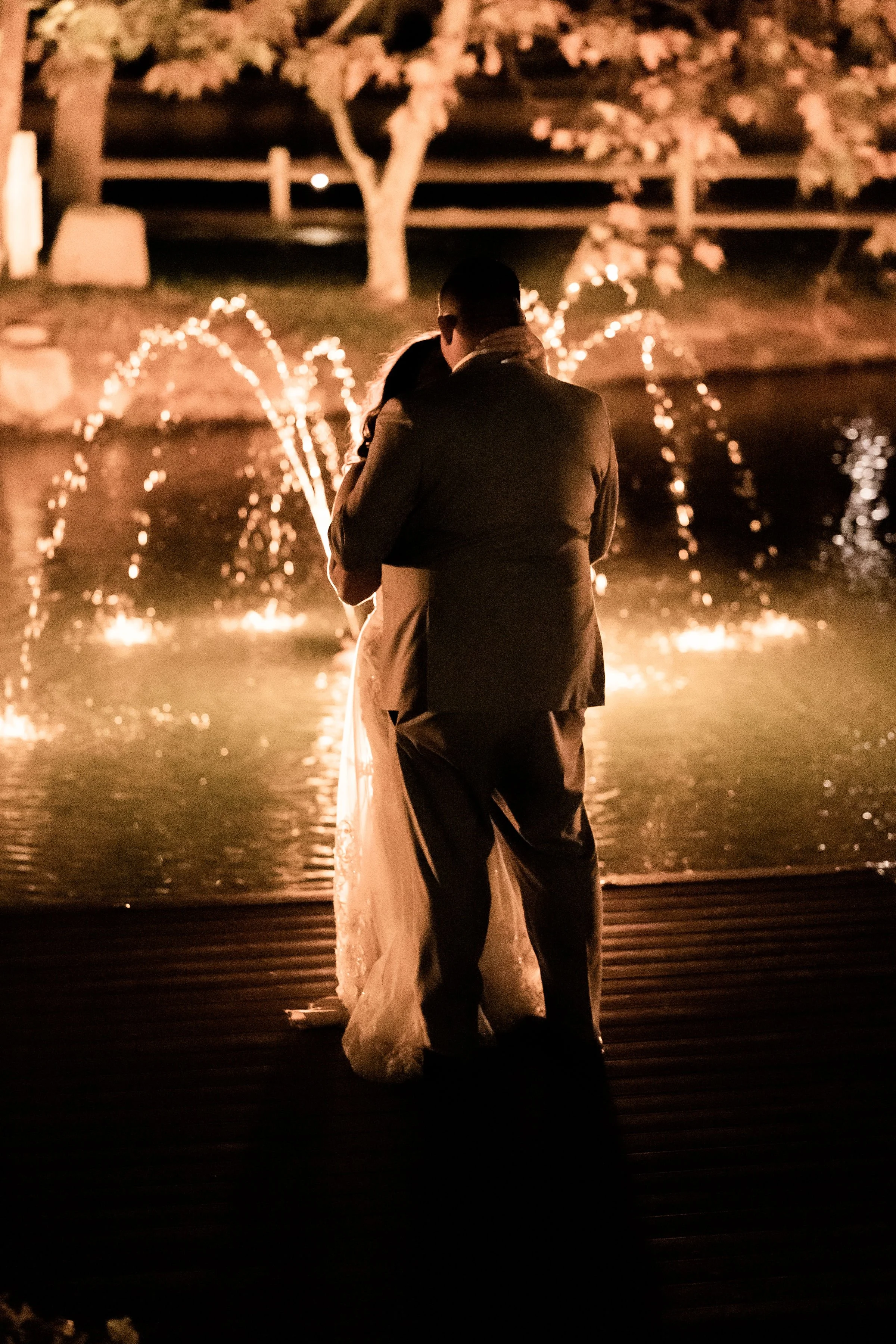 A couple in wedding attire embracing near a fountain with water jets illuminated at night.
