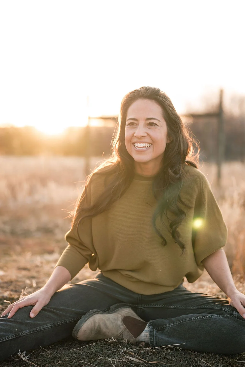 A young woman with long dark hair, wearing a brownish-green sweatshirt, sitting cross-legged outdoors on dry grass during sunset, smiling happily.