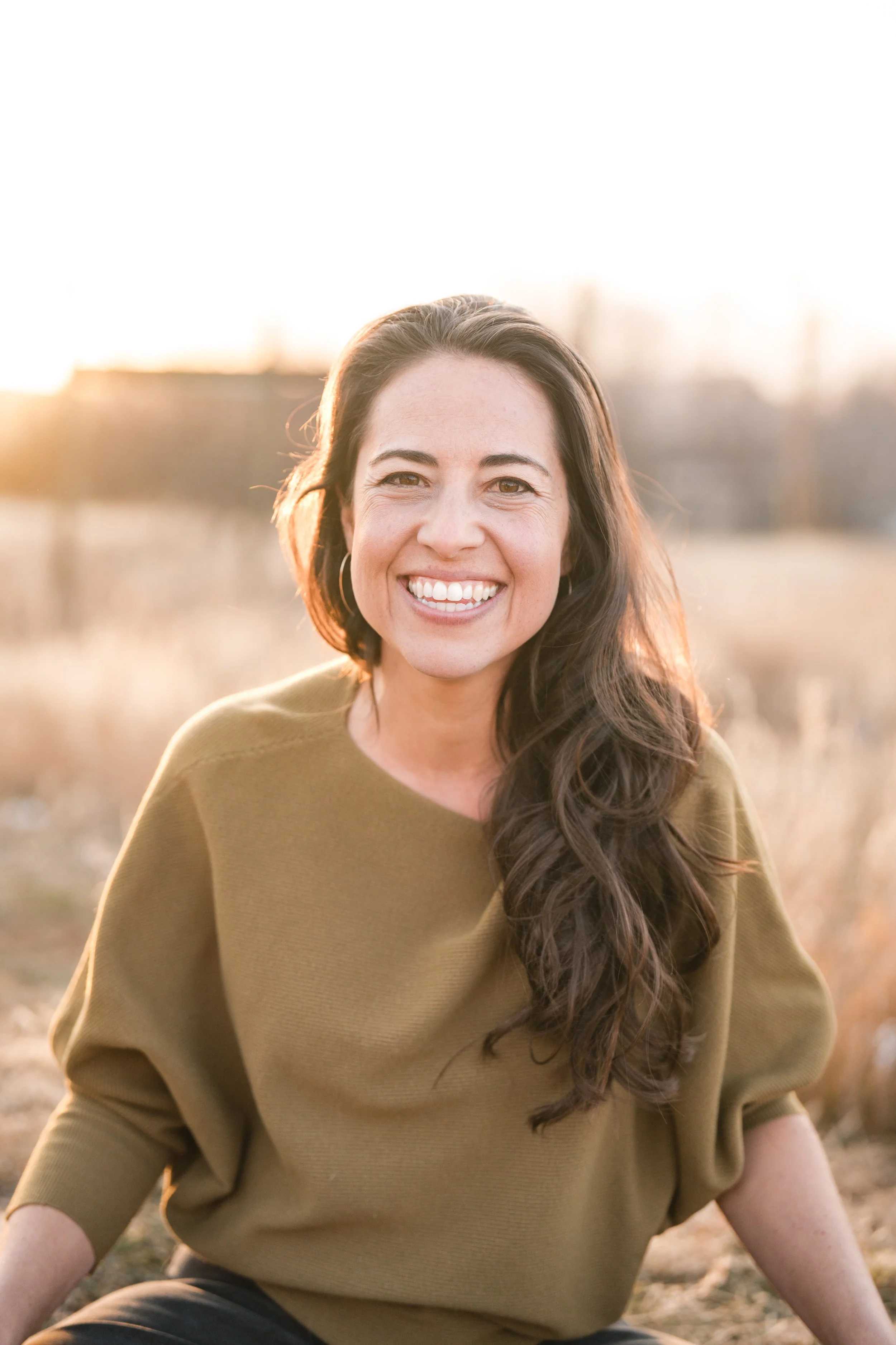 A woman with long dark hair and a wide smile, sitting outdoors during sunset in a field.