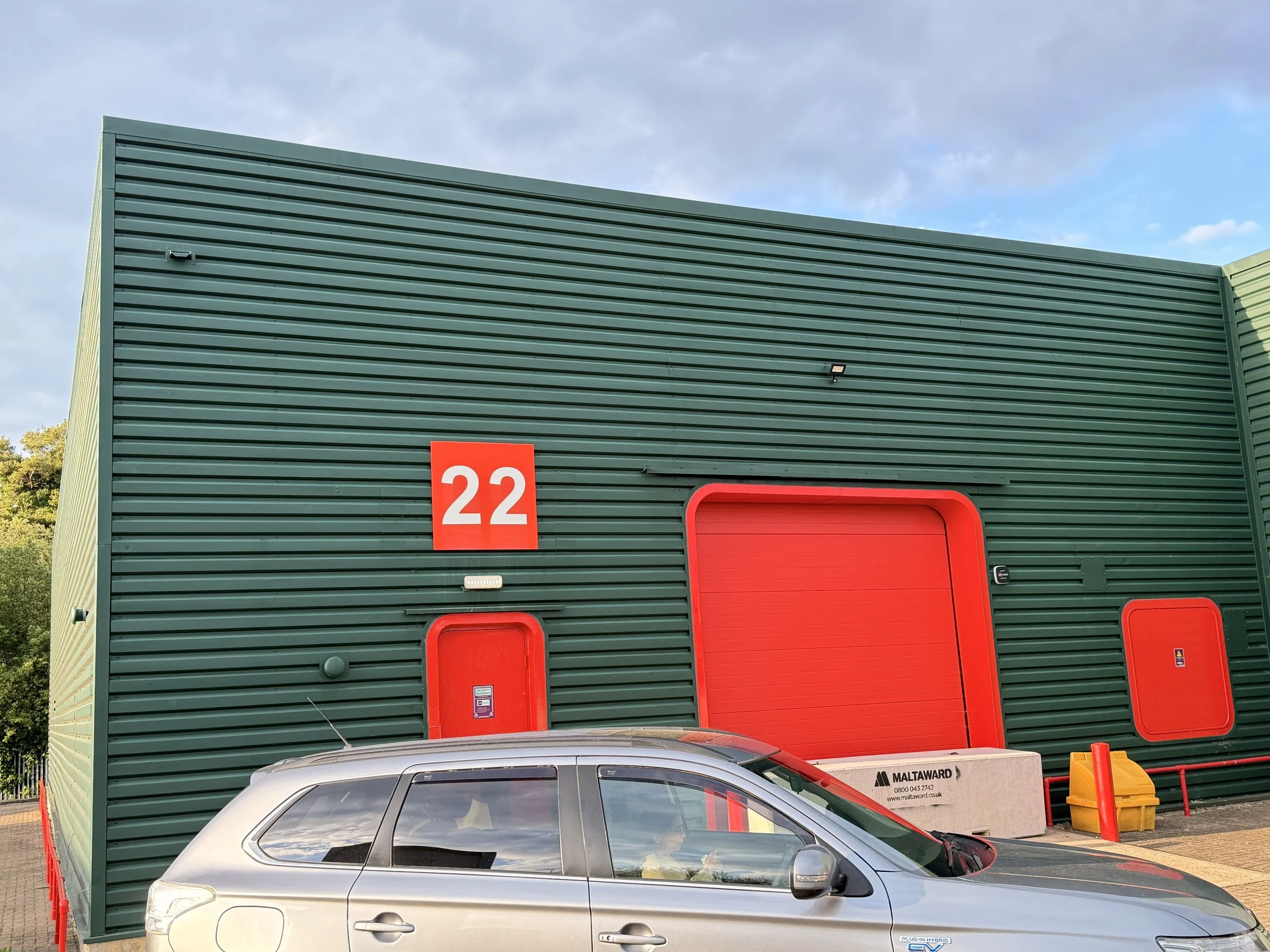 A green industrial building with a large red door and a red number 22 sign. A partial view of a silver car in the parking lot in front of the building.