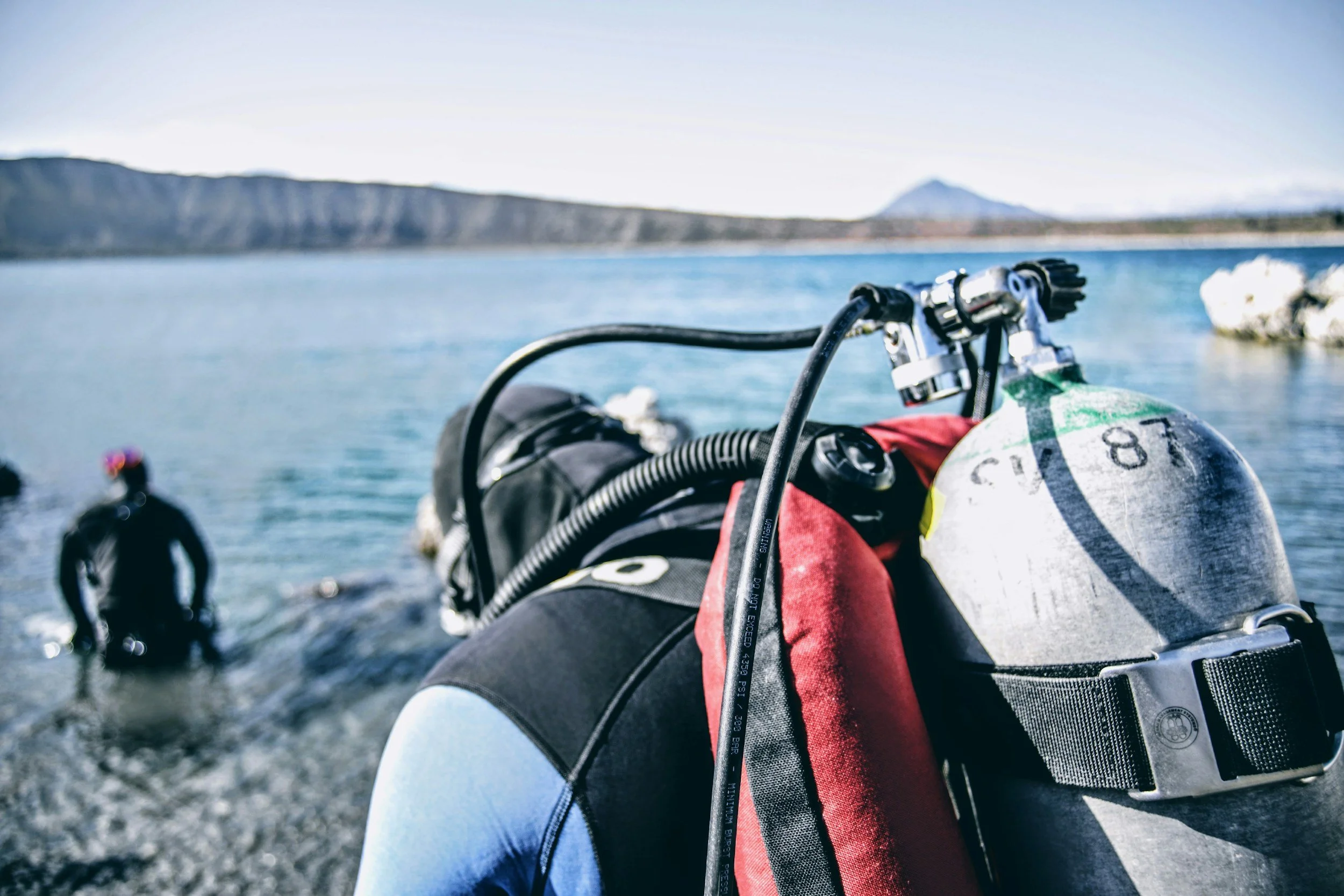Person in wetsuit with scuba gear preparing on a shore next to the water, with a diver swimming nearby and mountains in the distance.