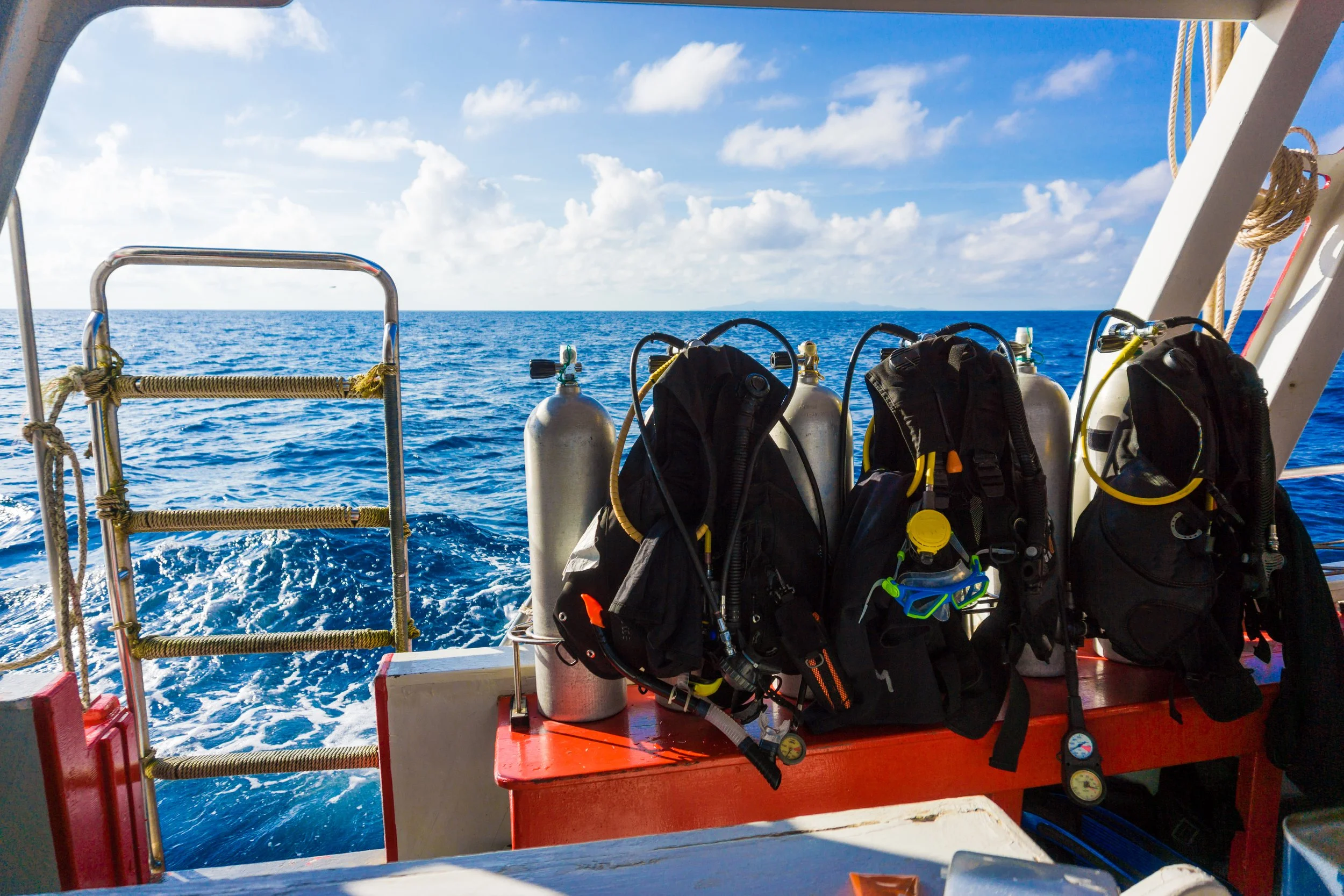 Scuba diving equipment including tanks, buoyancy compensators, and masks left on the deck of a boat with the ocean and sky in the background.