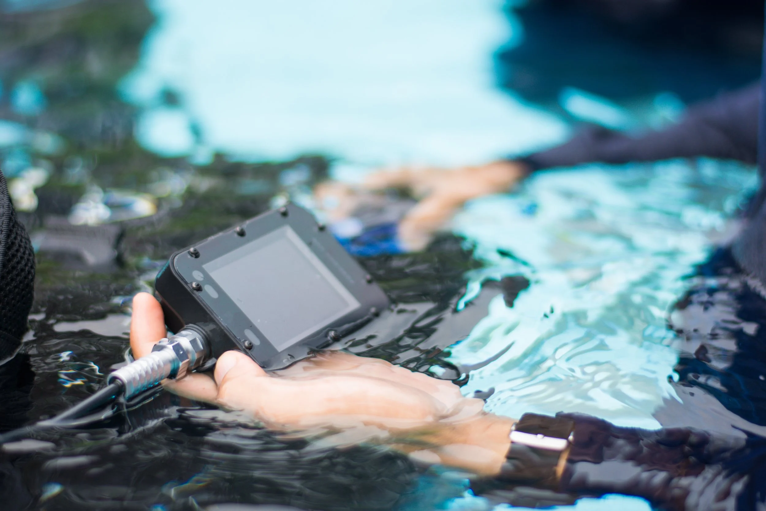 A diver holding a scuba console with a screen and a silver connector, partially submerged in water during a diving or underwater exercise.
