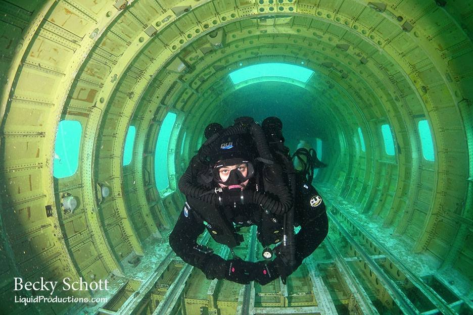 An underwater diver in black gear and mask inside a yellow cylindrical structure with windows, looking at the camera.