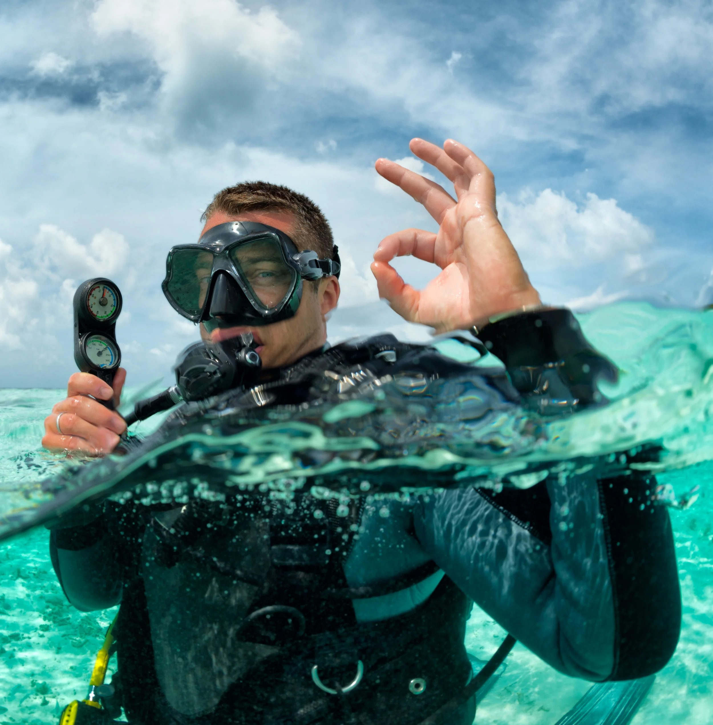 A scuba diver in water holding a dive gauge, making an OK hand signal with his other hand, wearing a diving mask and wetsuit under a cloudy sky.