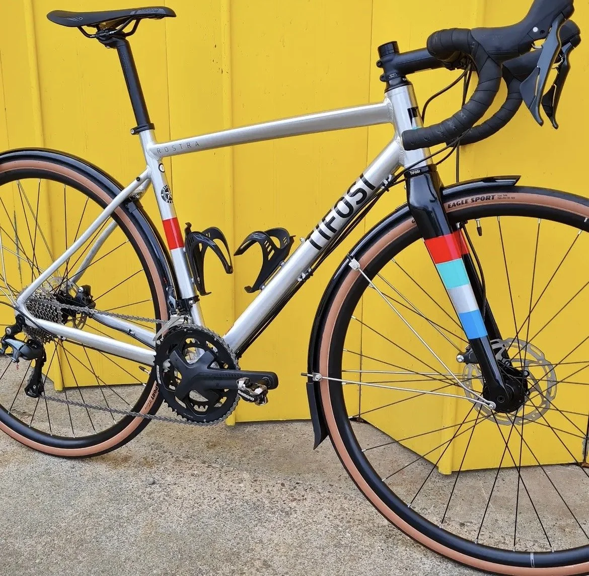 A Tifosi modern Rousta gravel or adventure bike with a silver frame and colorful accents, black handlebars, and tan-colored tires, leaning against a yellow wall.