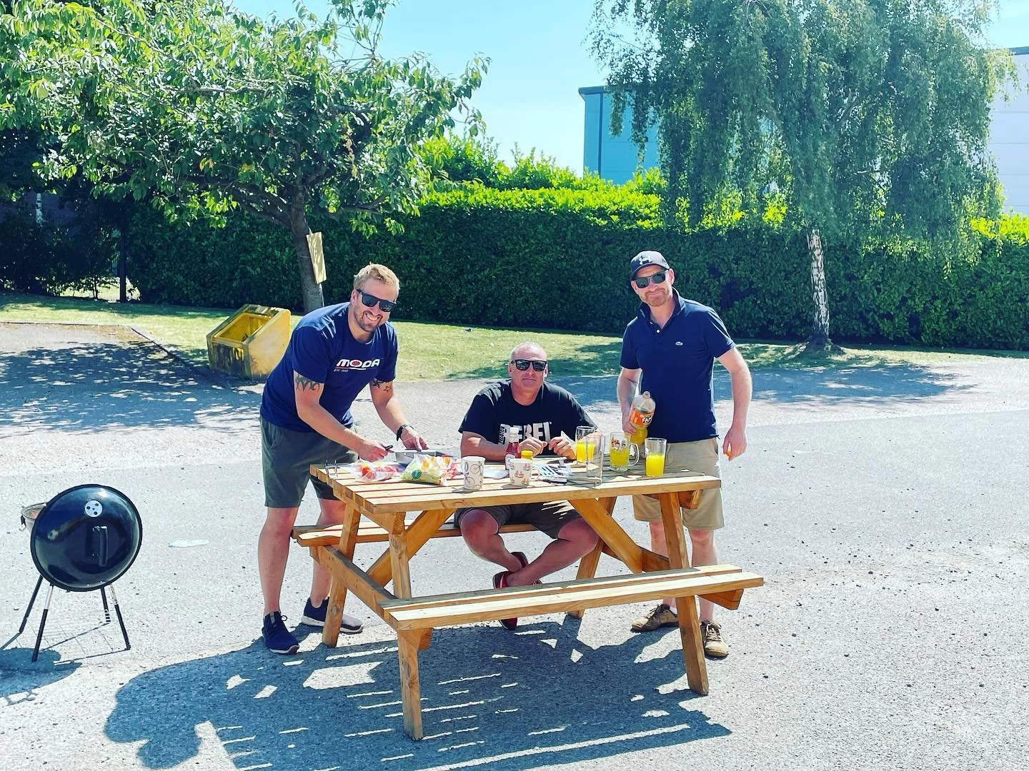 Three men are gathered around a wooden table outdoors on a sunny day. One is standing on the left, wearing sunglasses, a blue T-shirt, and shorts, preparing food. The man in the middle is sitting at the table, wearing sunglasses and a black T-shirt, 