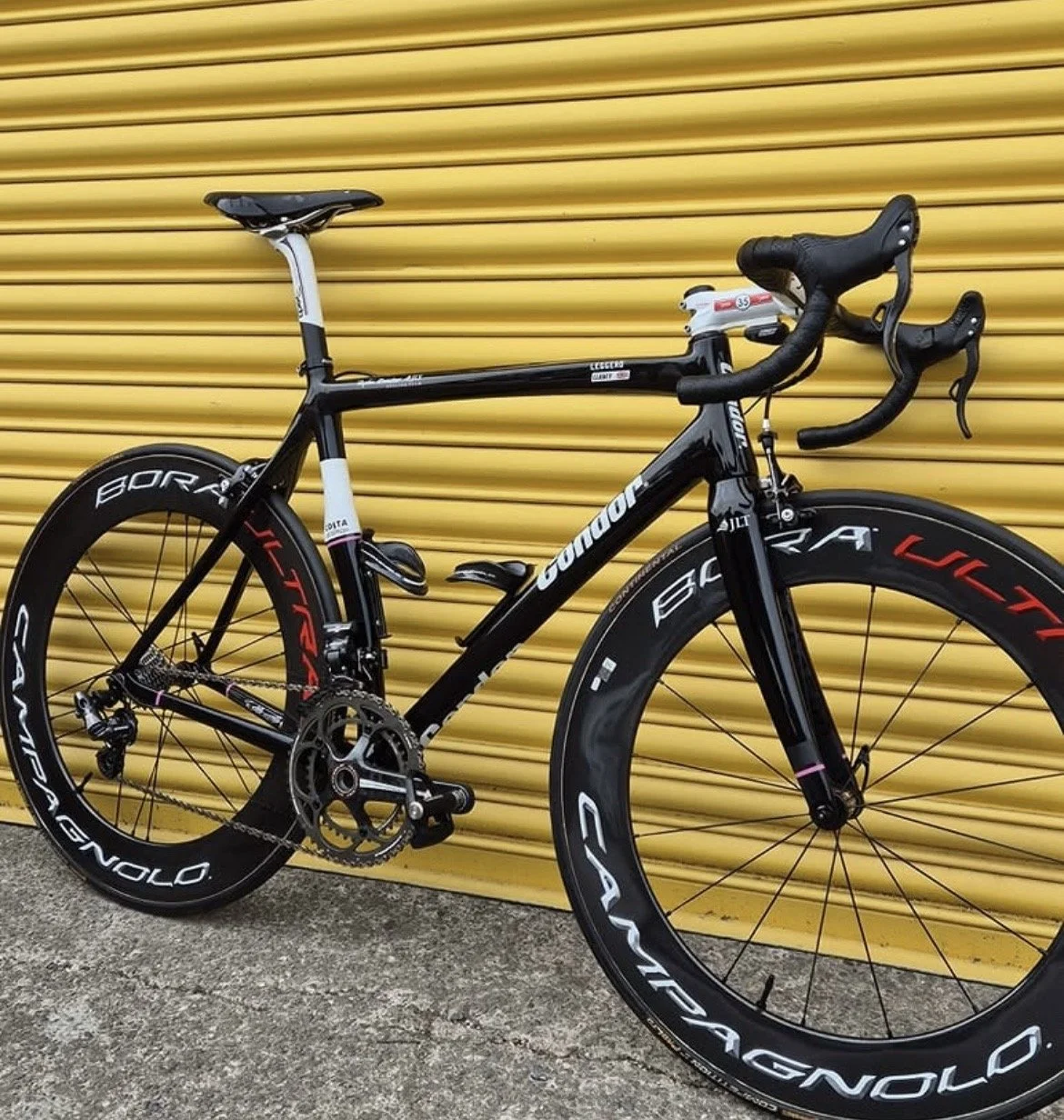 A black and white Cannondale road bicycle with deepdish carbon wheels, silver and red accents, and electronic gear shifters, positioned against a yellow corrugated metal wall.