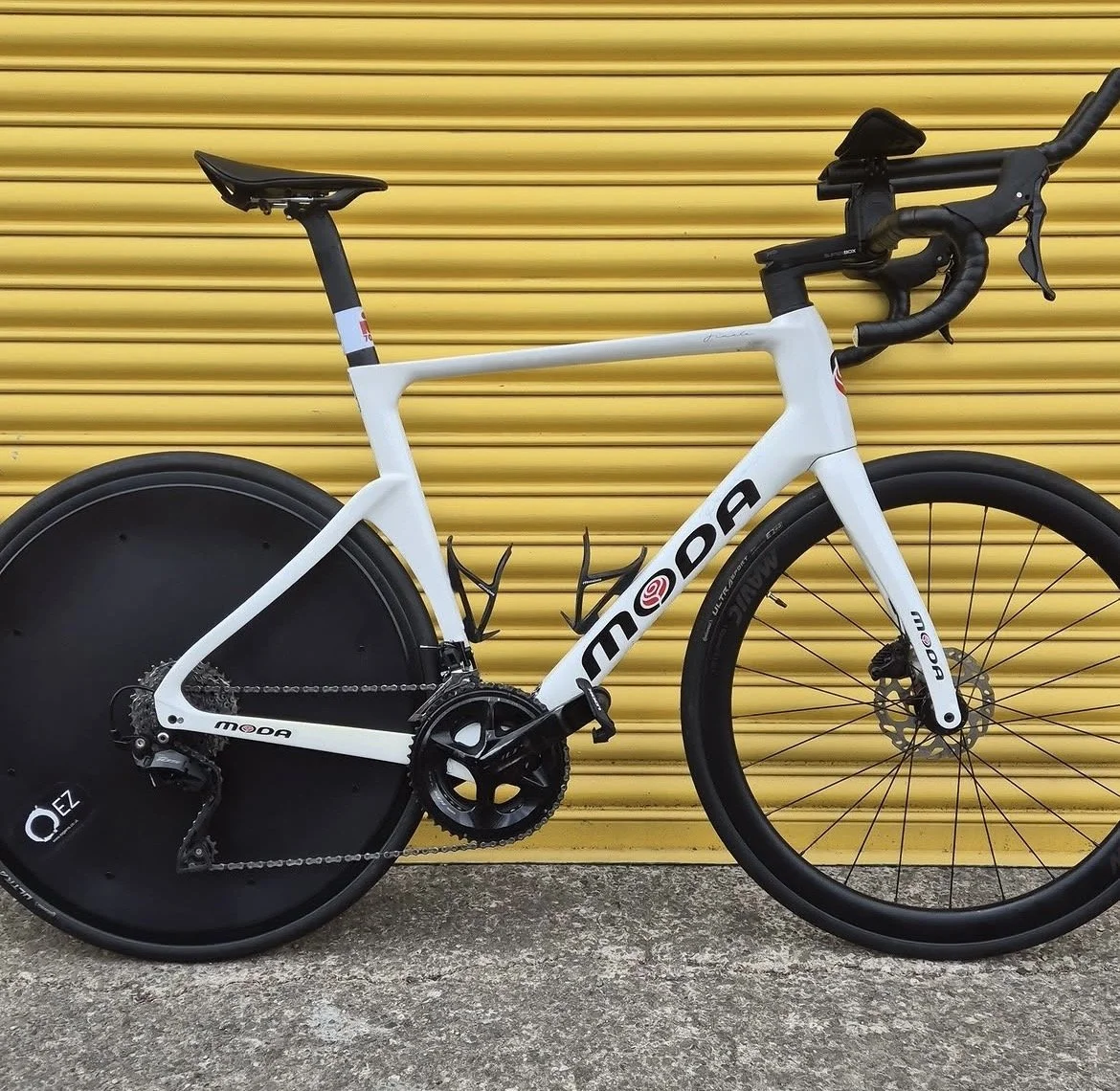 A white modern road bicycle with black tires and a black disc rear wheel, leaning against a yellow metal roll-up door.
