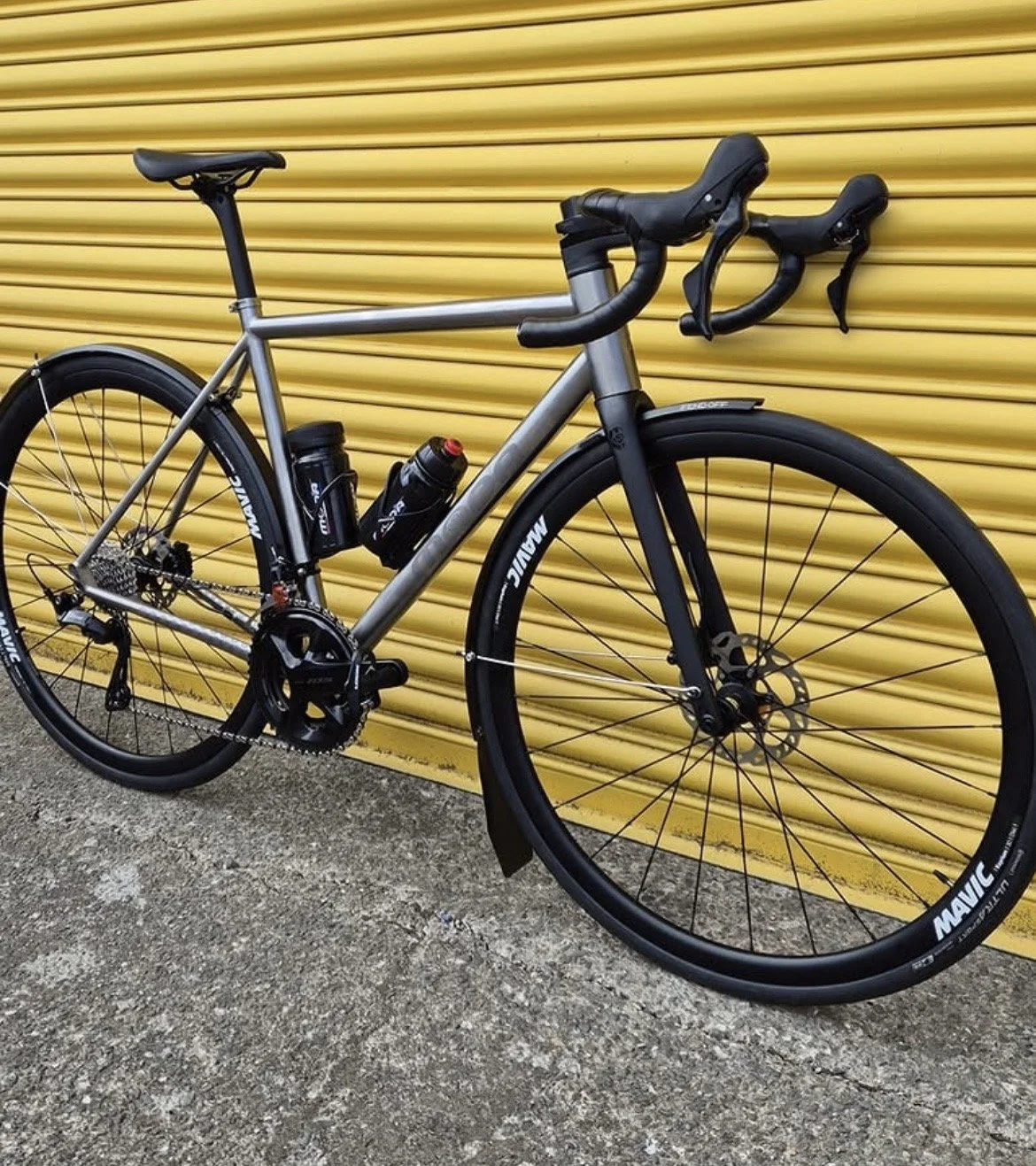 A silver and black road bicycle with drop handlebars, water bottles, and disc brakes, parked against a yellow corrugated metal wall.