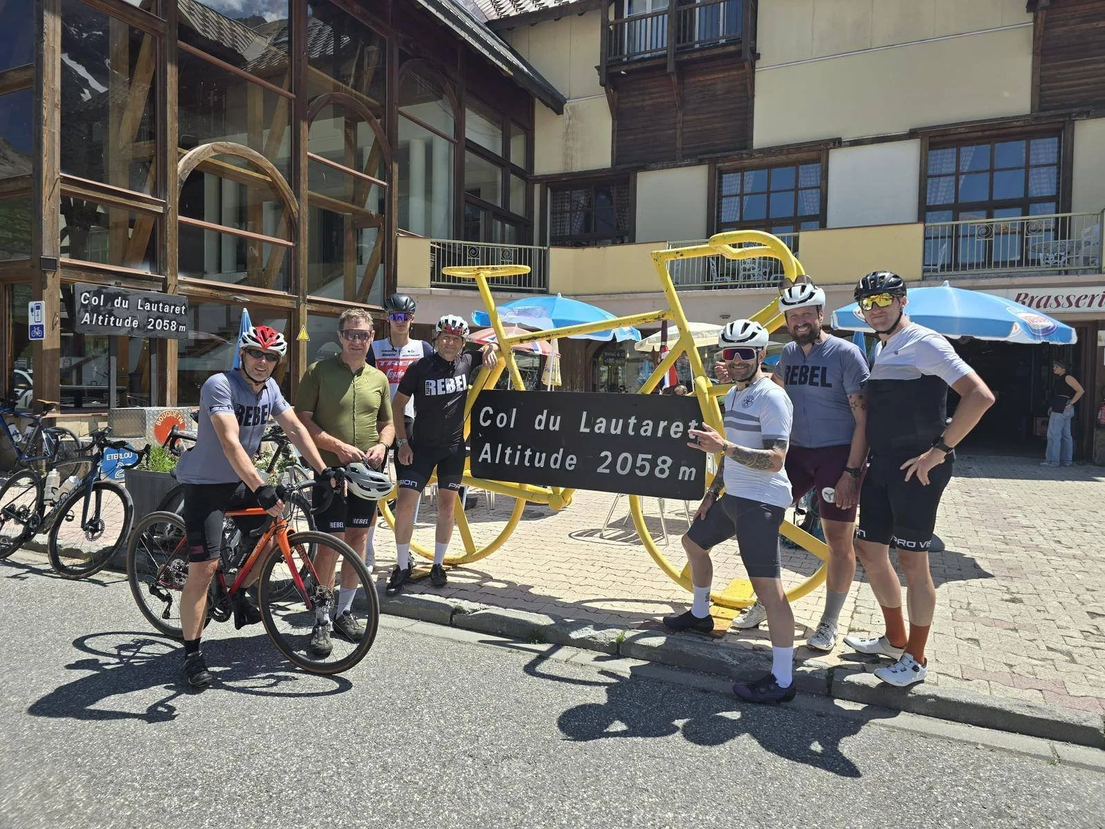 Group of seven cyclists in front of a building on a sunny day. They are standing next to outdoor bike racks and a yellow bike-shaped sculpture that displays a sign reading "Col du Lautaret Altitude 2058 m." The cyclists are wearing helmets and sporty
