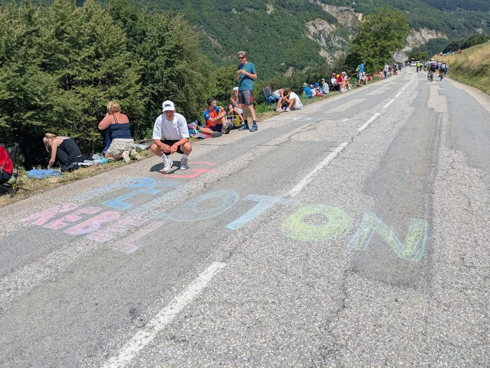 People sitting and standing along the side of a mountain road with colorful chalk messages on the pavement that say "Love" and "Hope" on a sunny day. Cyclists are riding in the distance, and the background features green trees and rocky cliffs.