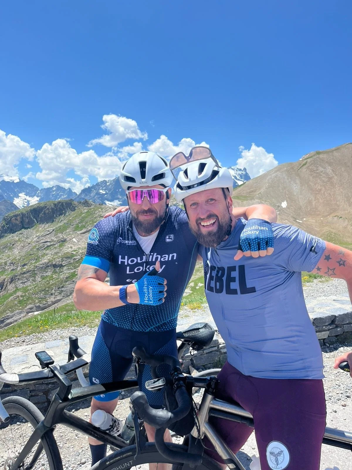 Two men in cycling gear and helmets smiling and posing with bikes in a mountain landscape under a blue sky with clouds.