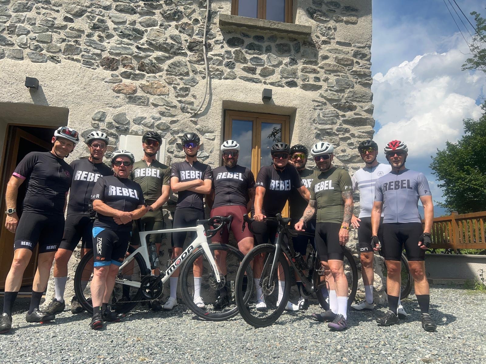 A group of eleven men in cycling gear standing outside in front of a stone building, some with bicycles, smiling at the camera on a sunny day.
