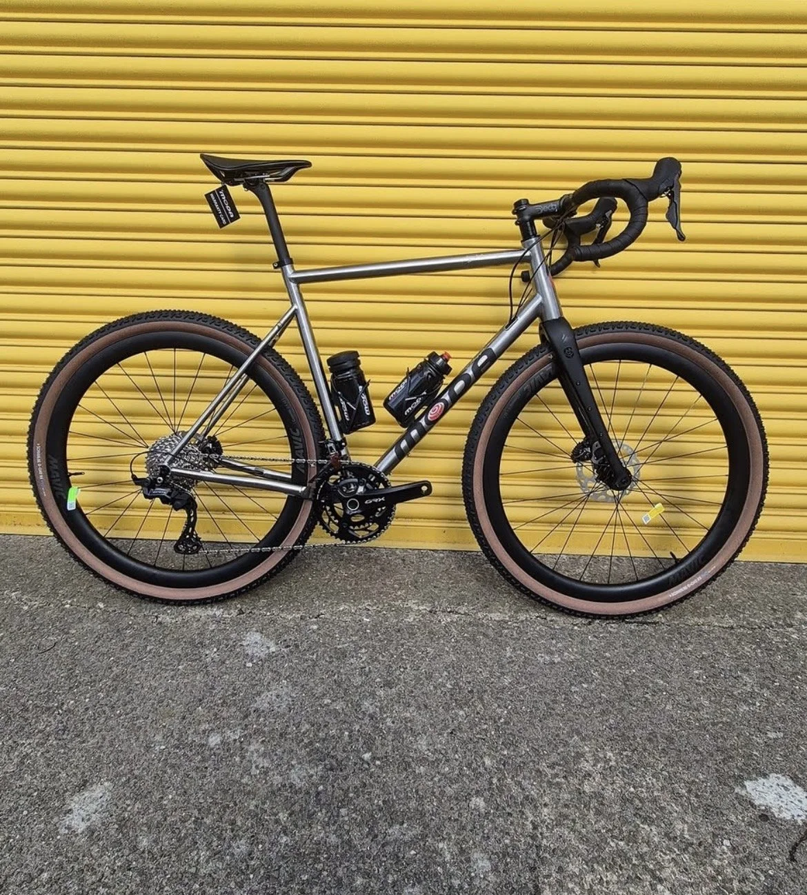A silver Trek mountain bike with black handlebars, tires with a tan sidewall, and pink accents, leaning against a yellow roll-up door on a concrete surface.