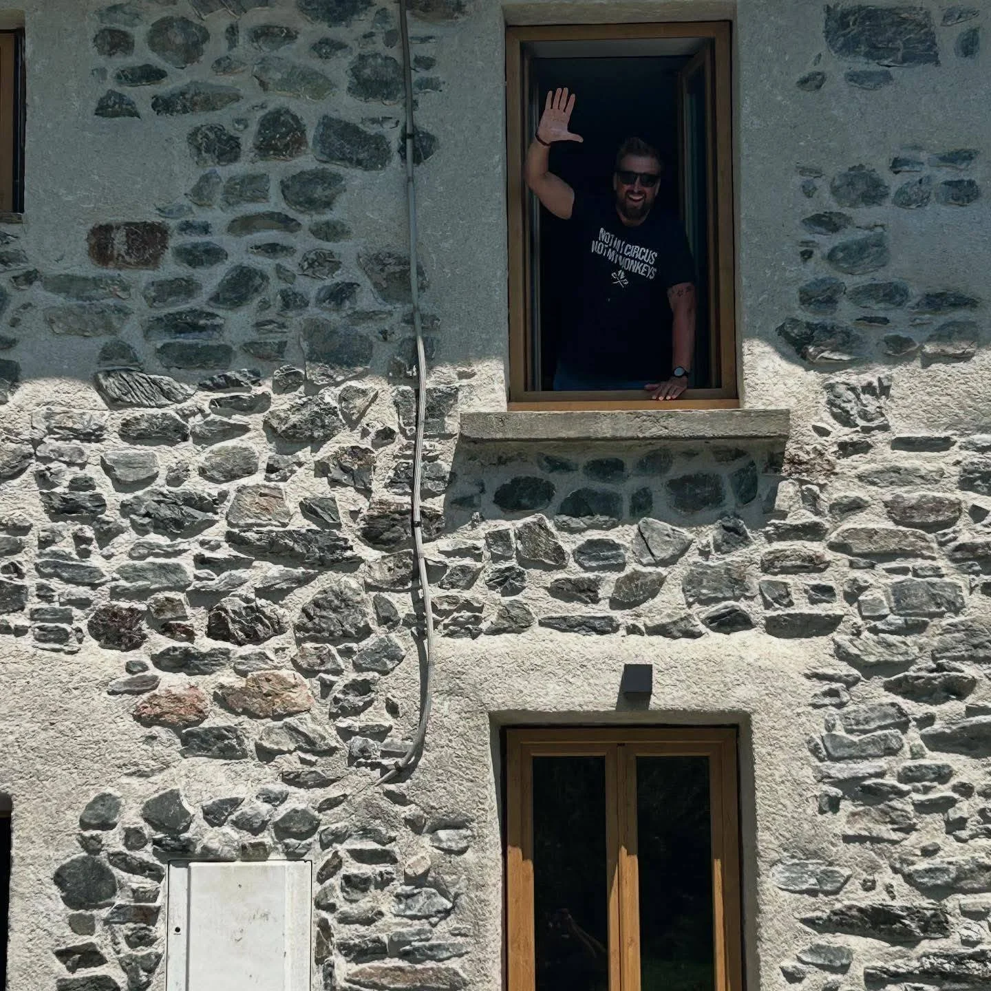 A man with dark hair, beard, sunglasses, wearing a black T-shirt waving from a mountain stone house window.