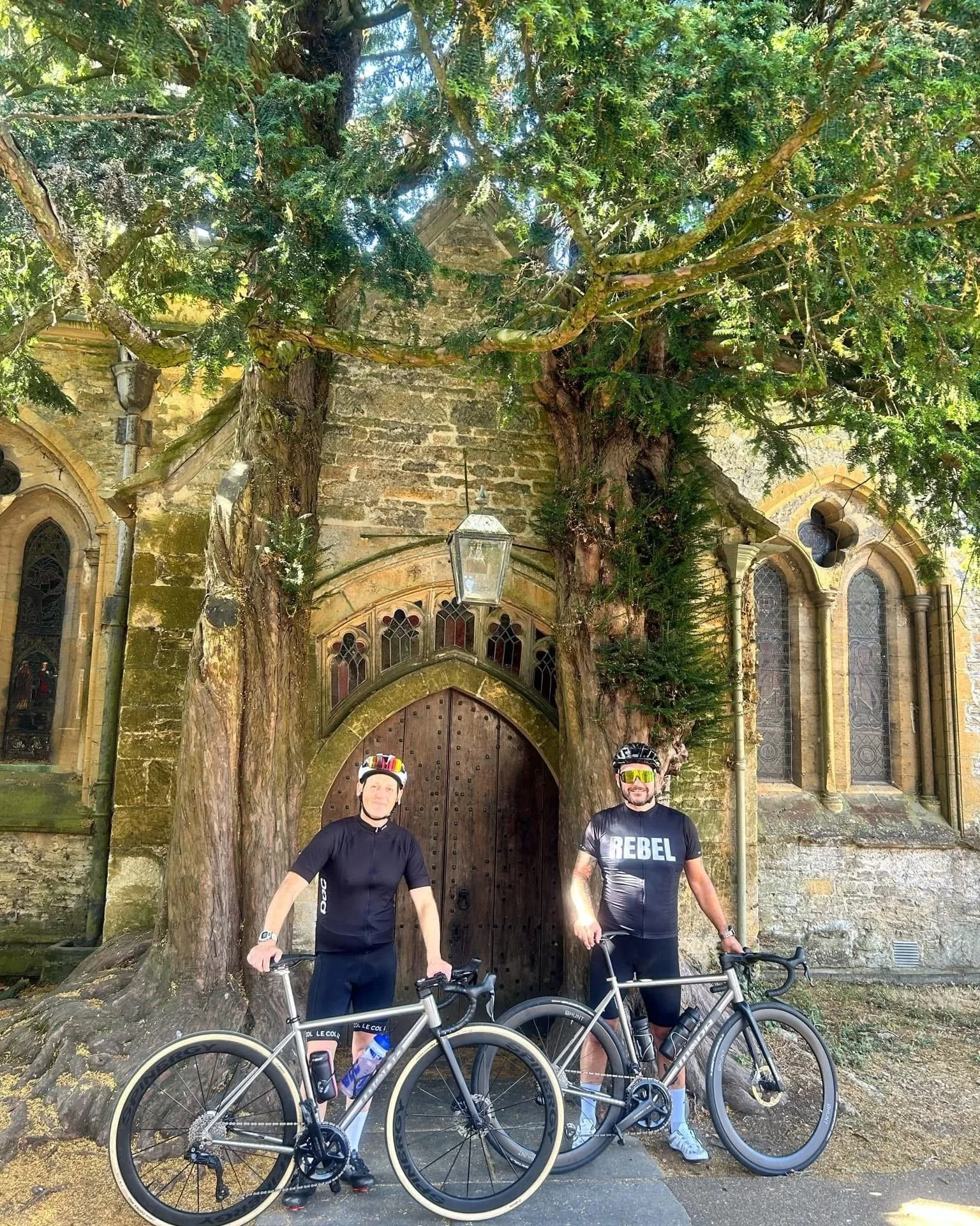 Two cyclists standing with their bikes in front of an old stone building with large arched windows, partially obscured by two large trees and a lamp post.