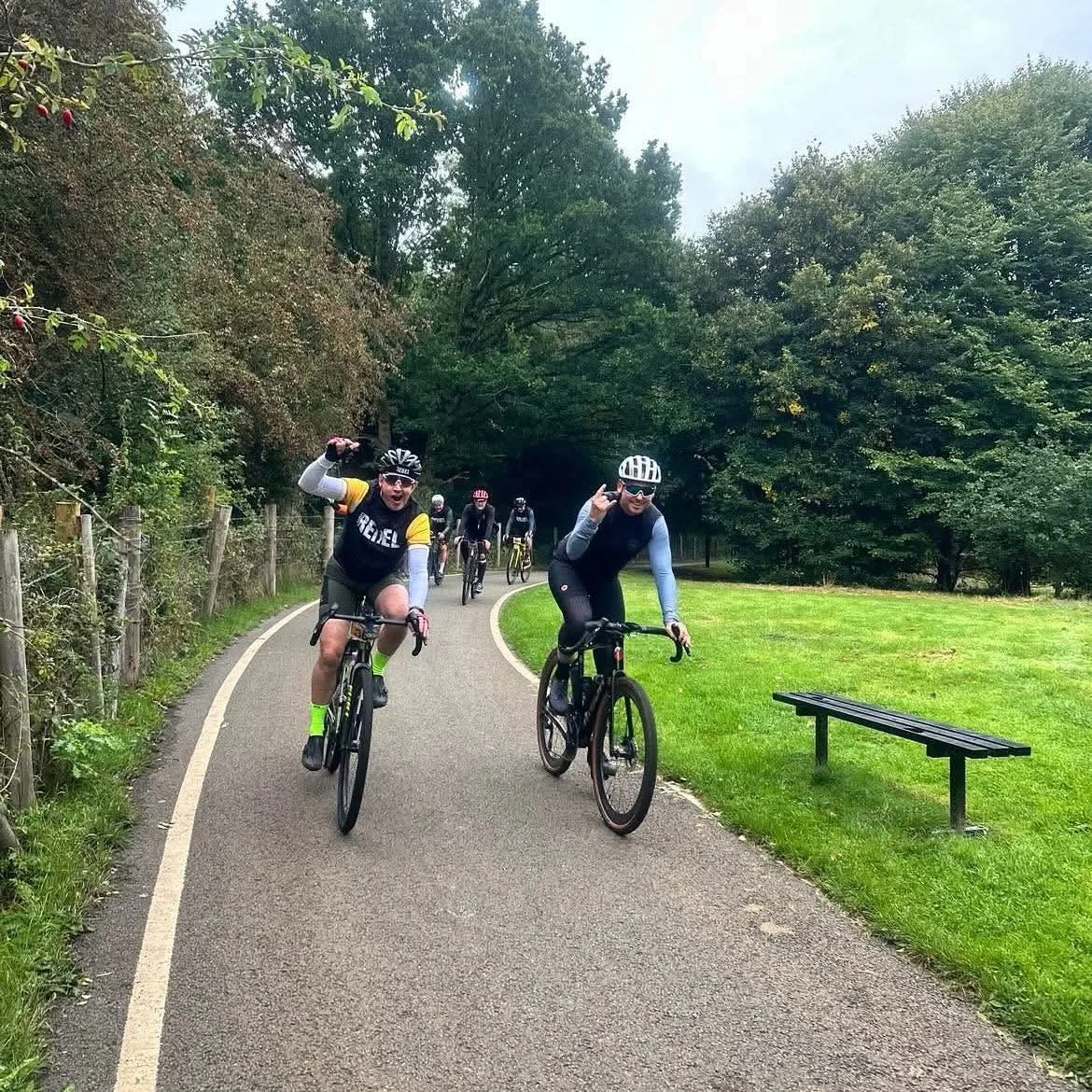 A group of cyclists riding on a paved trail through a park with green grass, trees, and a wooden fence on the side. Two cyclists are in the foreground, one making a peace sign and the other flexing his arm, smiling at the camera.