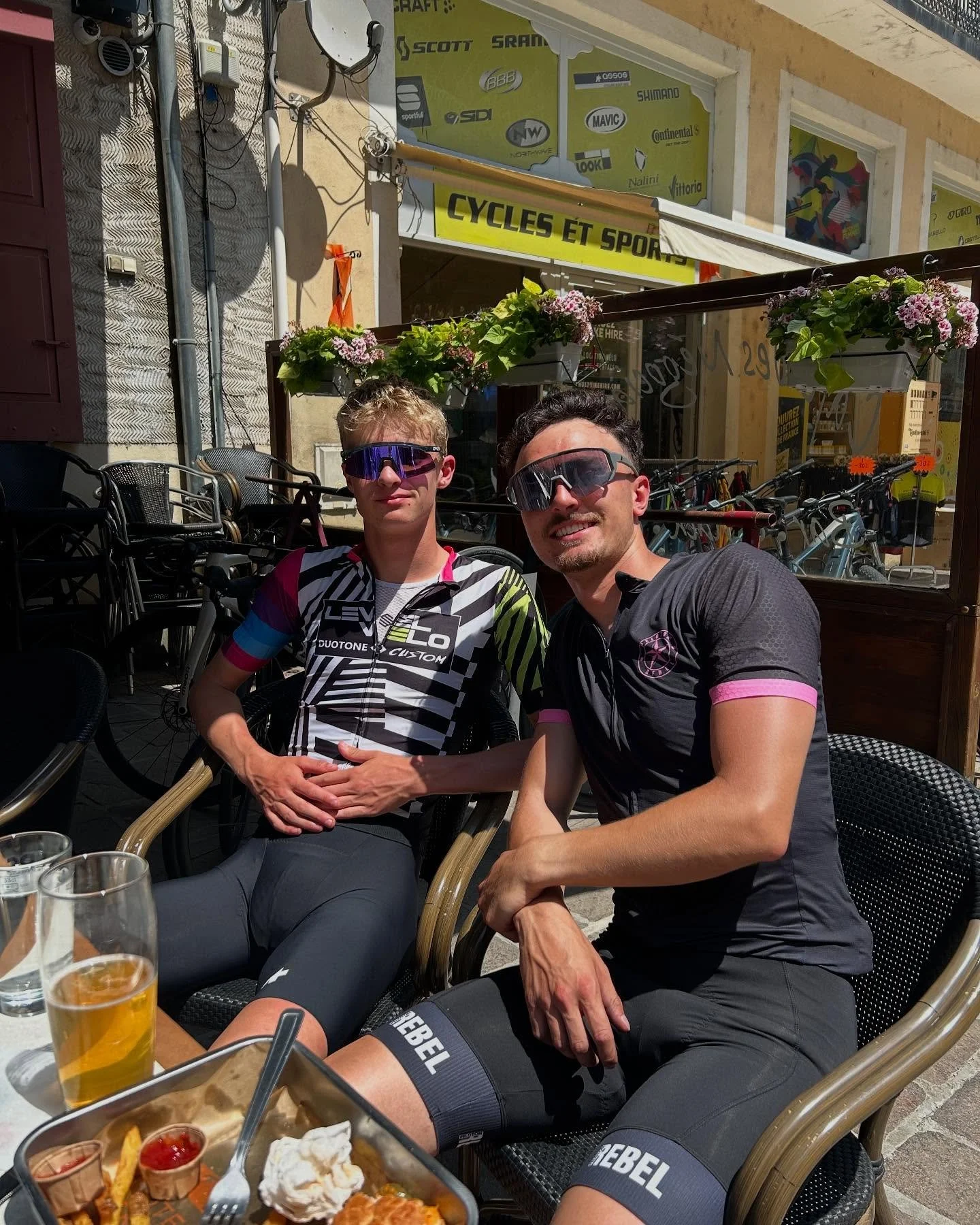 Two men sitting at an outdoor cafe table, wearing cycling jerseys and sunglasses, smiling at the camera. There are glasses of beer and a plate of food in front of them. Behind them, bicycles are parked, and a bicycle shop sign is visible on the build