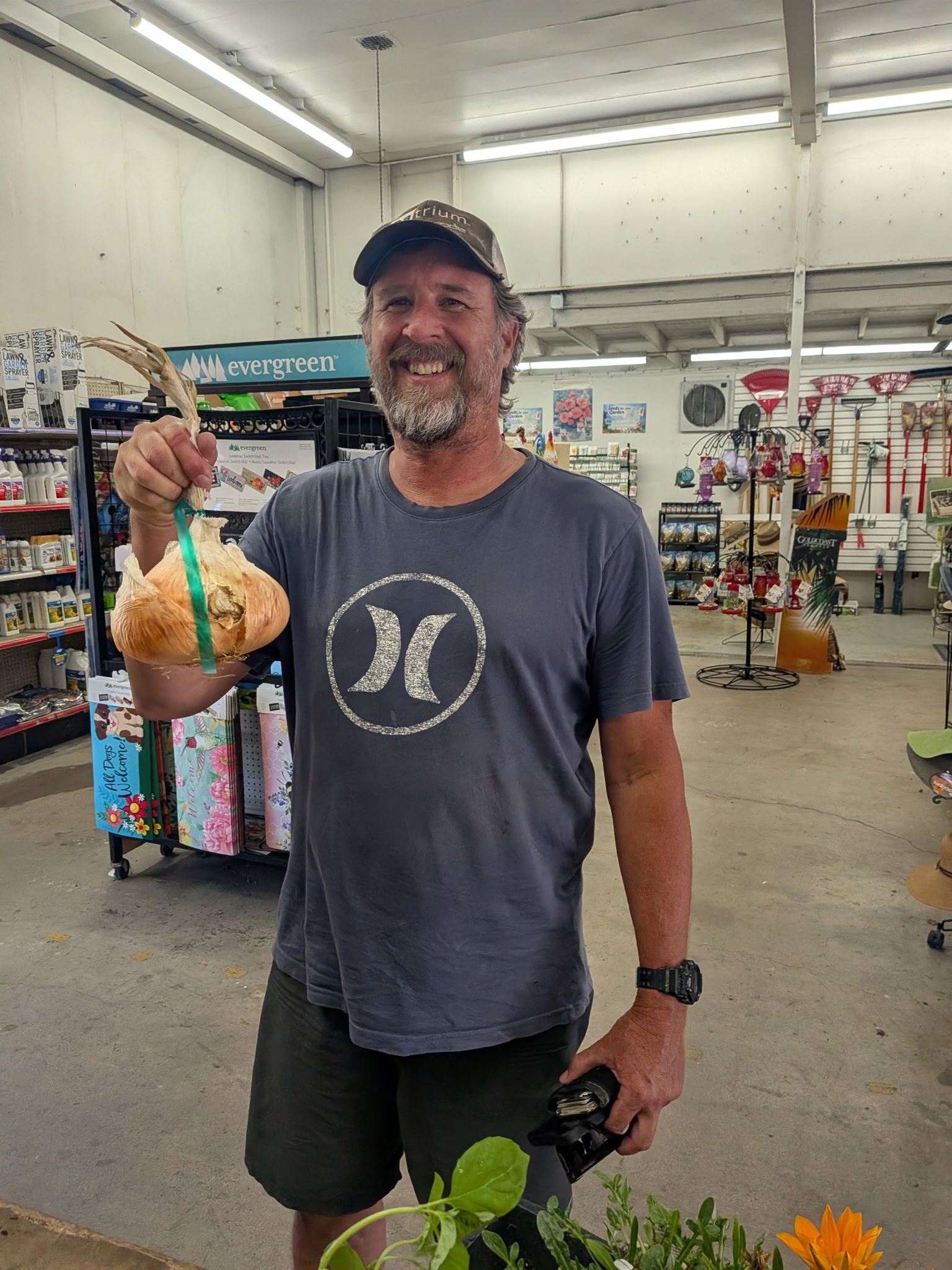 A smiling man in a dark gray t-shirt and black shorts is standing inside a store, holding a large peeled onion in his left hand and a remote control or electronic device in his right hand. The store has gardening products and decorations on the shelves and walls.