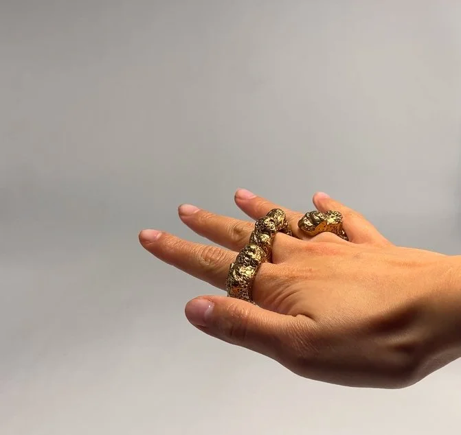 A hand with gold jewelry including a snake-shaped ring and a textured band ring set against a plain background.