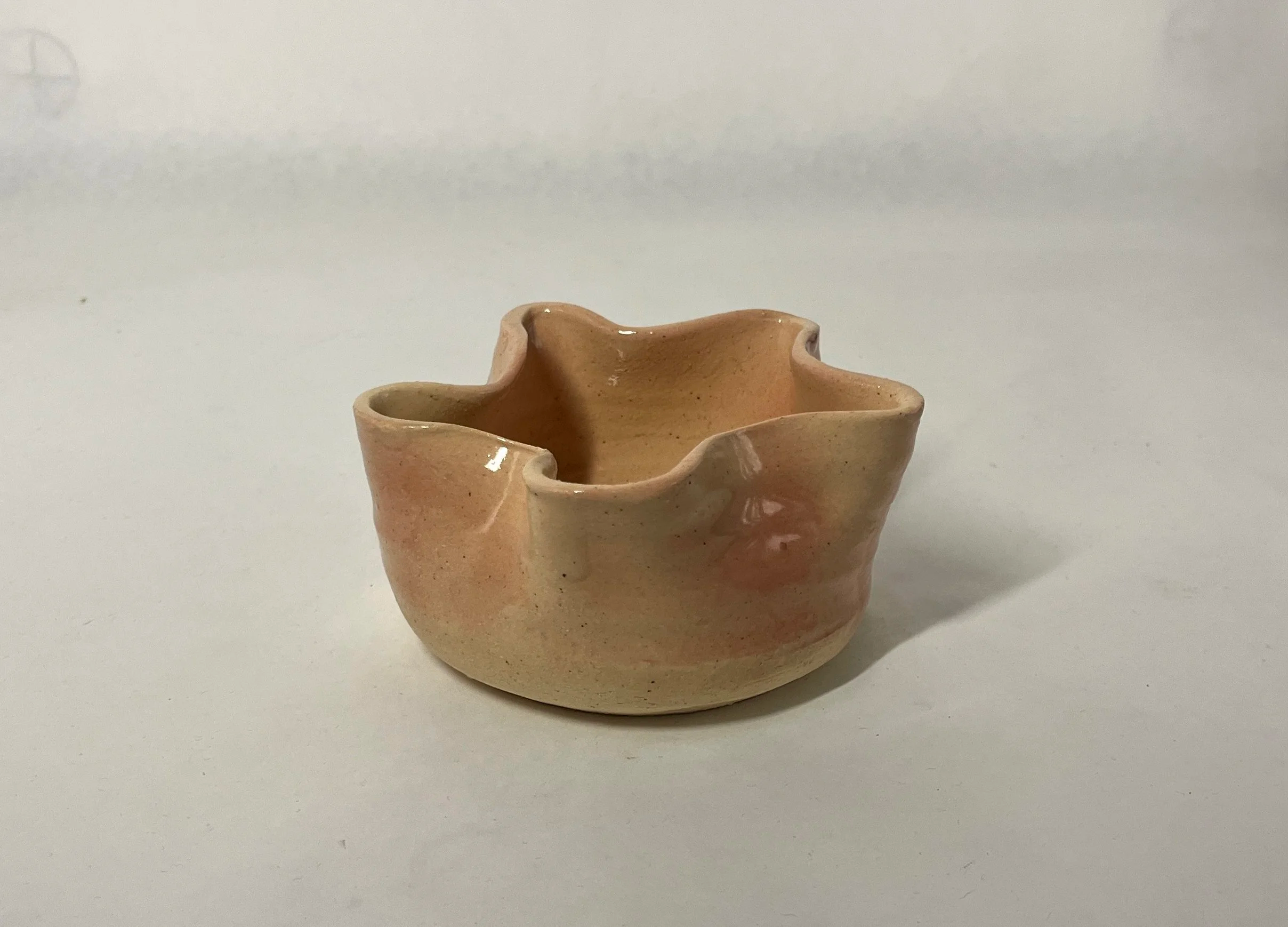 Small, irregularly shaped ceramic bowl with a wavy, uneven rim, glazed in soft peach and beige tones, placed on a plain white surface.
