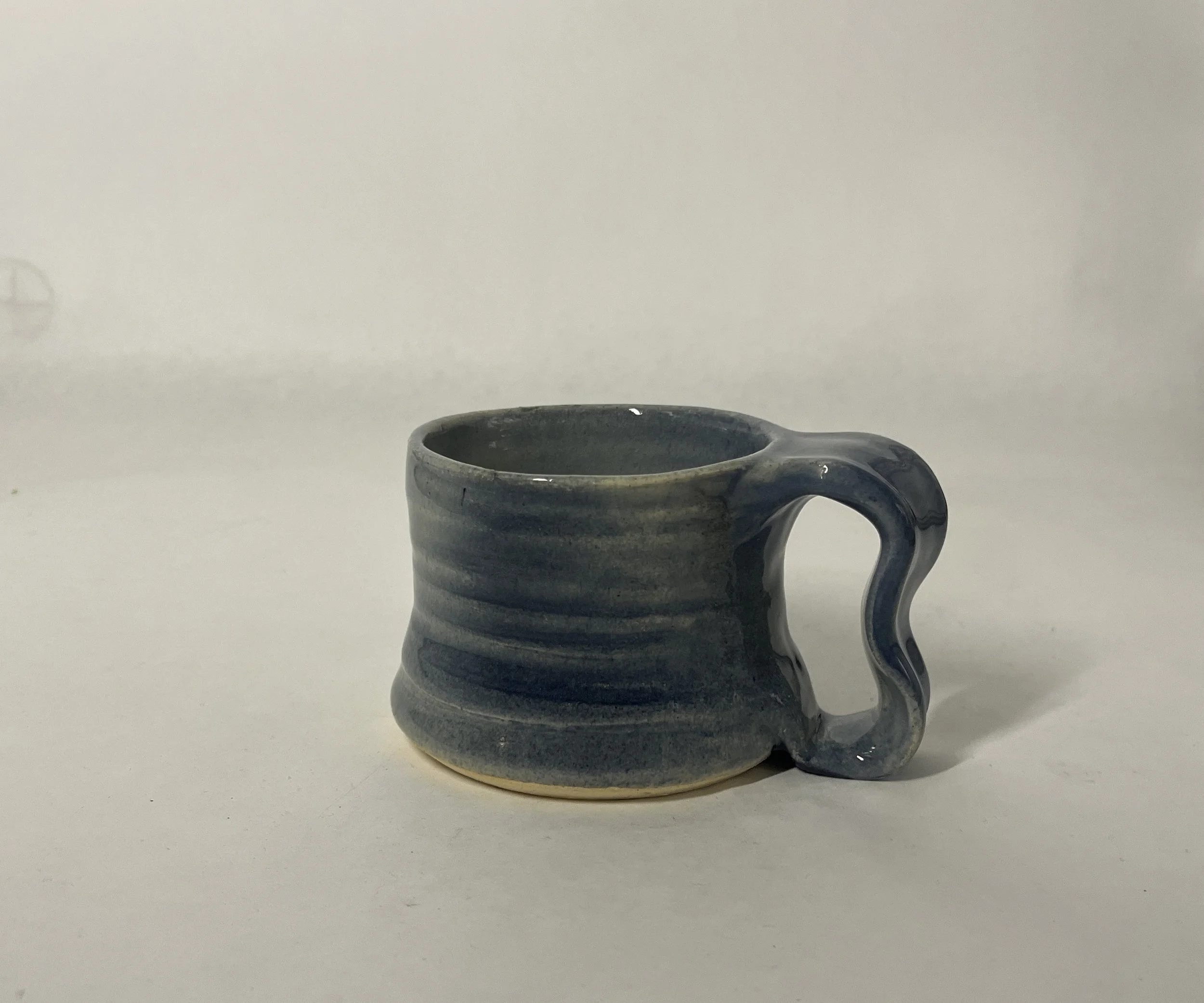 A small handmade ceramic mug with blue and gray stripes, with a curvy handle, placed on a white surface.