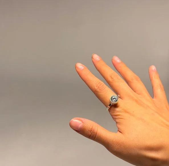 A hand with a large ring on the ring finger, featuring a round gemstone, against a plain gray background.